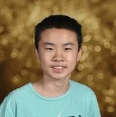 A young boy with short black hair smiling, wearing a light blue shirt against a gold blurred background.