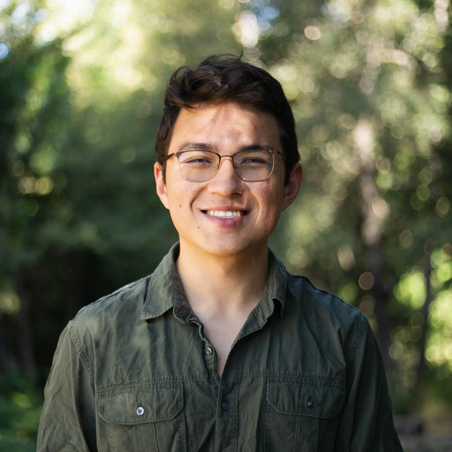 Close-up of a young man smiling, wearing glasses and a green button-up shirt, outdoors with blurred green foliage background.
