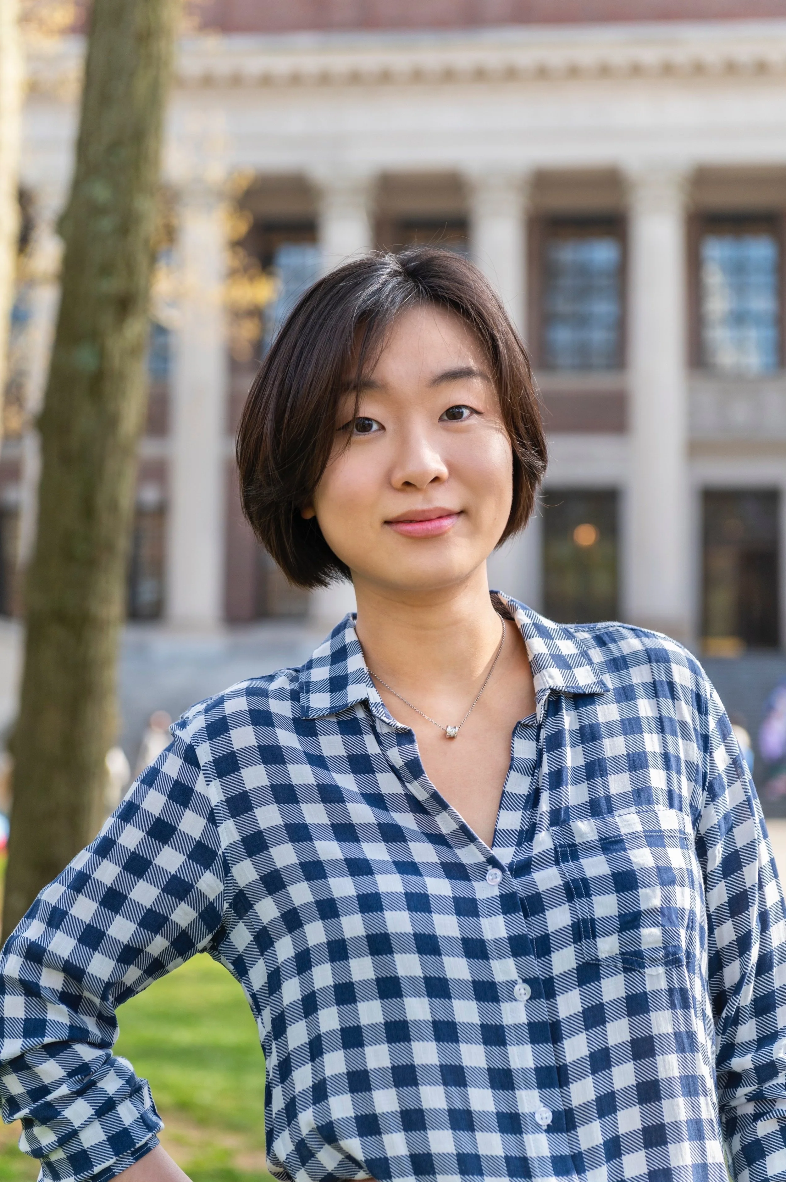 A woman with short black hair and a checkered blue and white shirt stands outdoors, smiling slightly at the camera, with a building and trees in the background.