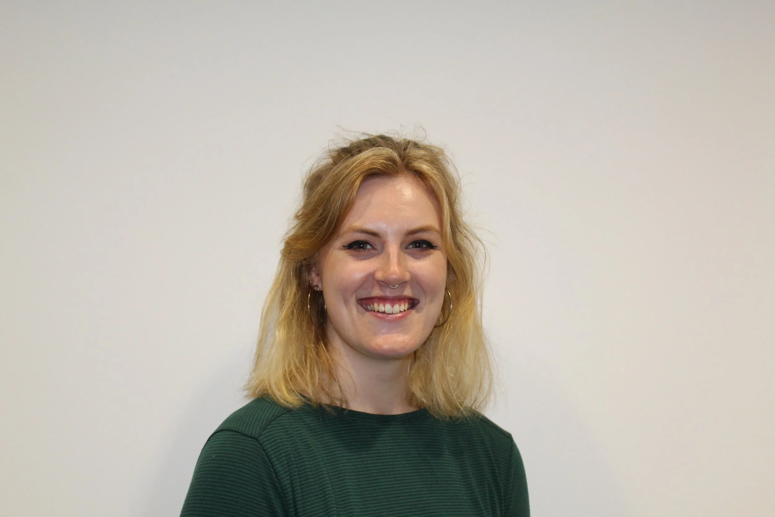 A smiling woman with blonde hair and hoop earrings, wearing a green striped shirt, standing against a plain white wall.