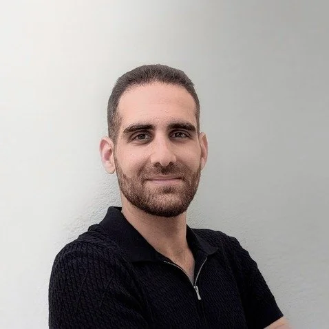 A young man with short dark hair and a beard, smiling, wearing a black zip-up sweater, standing against a plain light-colored wall.