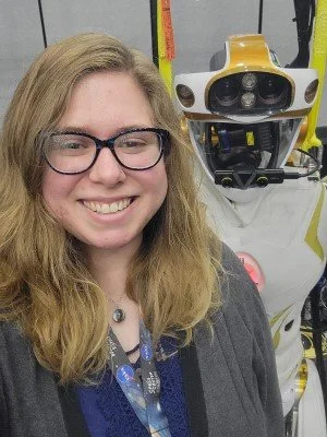 A woman with glasses smiling for a photo next to a humanoid robot in a laboratory or technology setting.