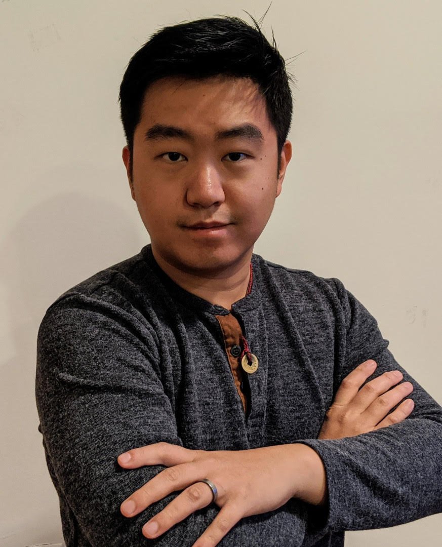 A young man with short black hair and light skin standing in front of a plain light-colored wall, crossing his arms and looking at the camera.