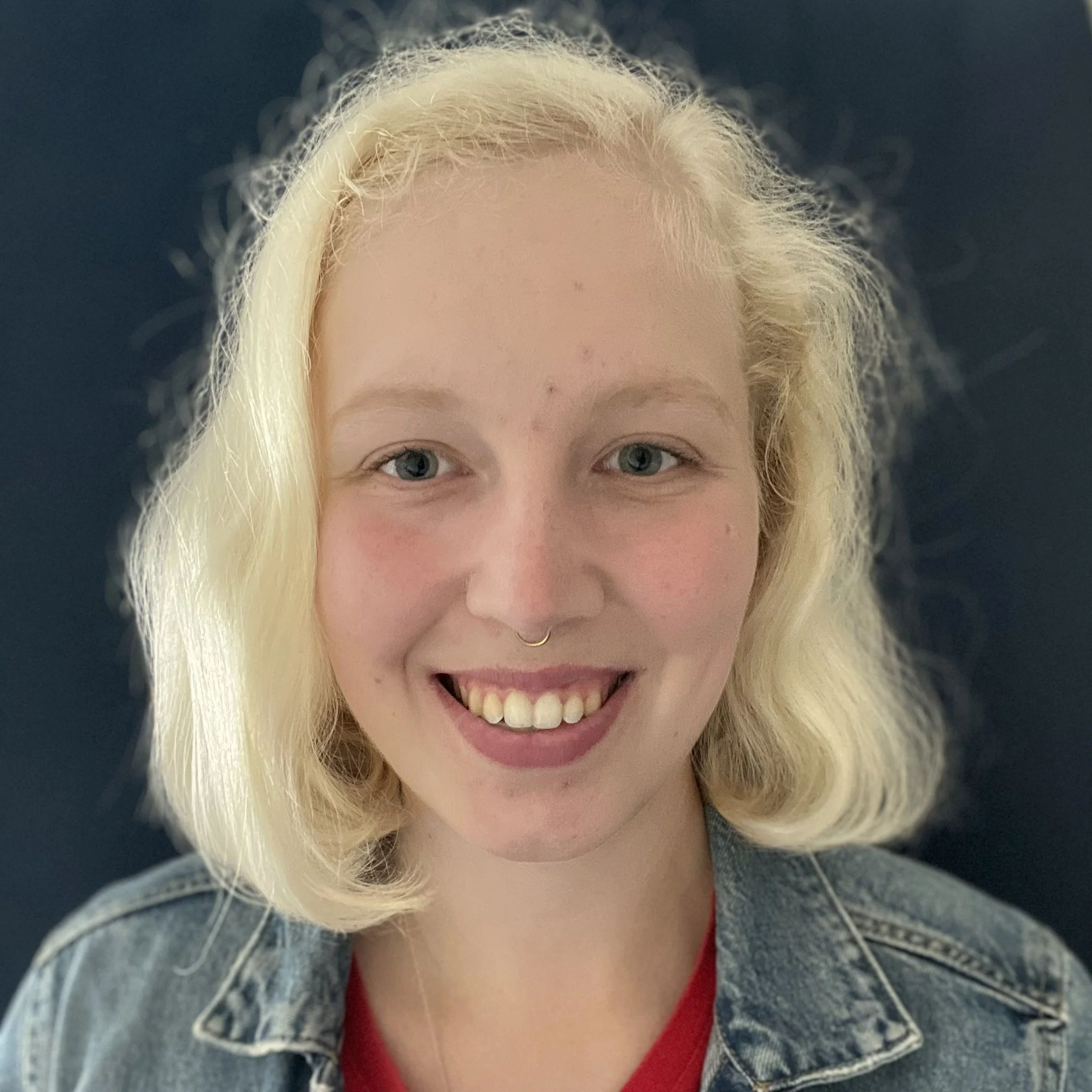 A smiling woman with blonde hair, wearing a necklace and a denim jacket, close-up shot with a dark background.