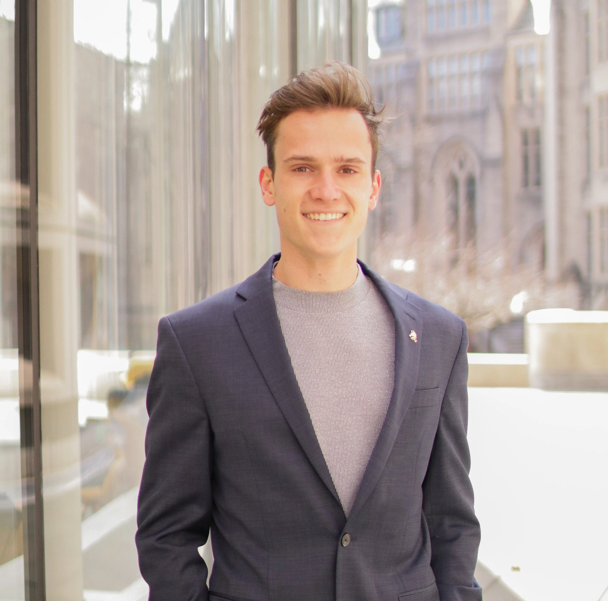 A young man with light skin, brown hair, and blue eyes smiling, standing in a modern building with large glass windows and an urban cityscape in the background. He is dressed in a dark blazer over a light-colored shirt.