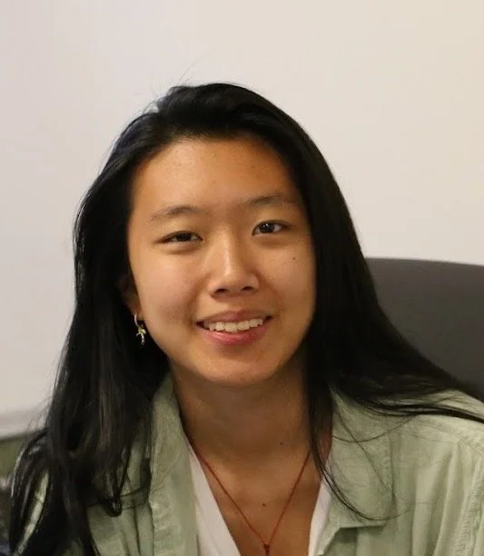 Young woman with long black hair smiling, wearing earrings and a green jacket, sitting indoors.