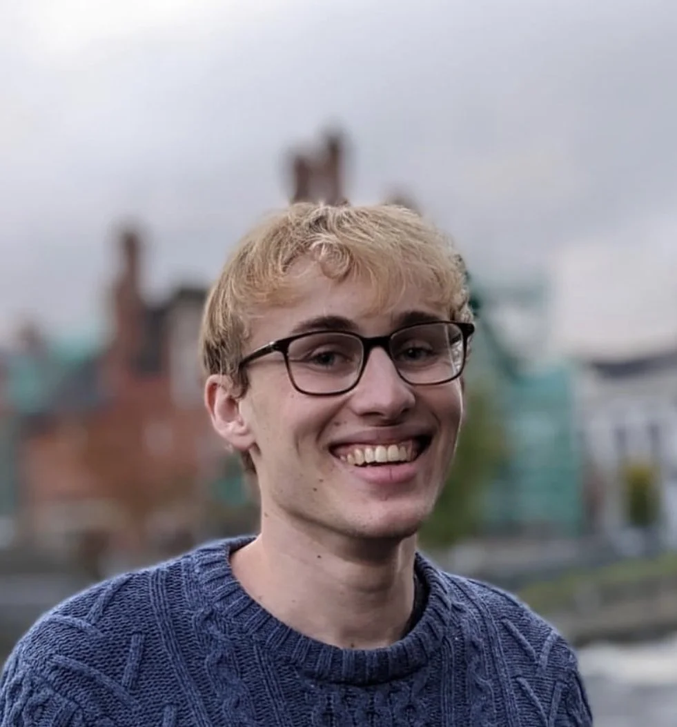 A young man with glasses and blonde curly hair smiling in an outdoor setting with buildings and cloudy sky in the background.