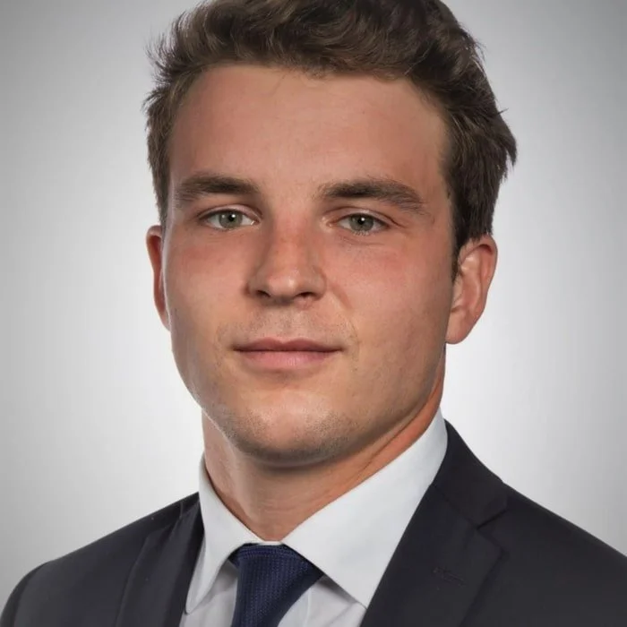 Professional headshot of a young man in a suit and tie against a plain gray background.