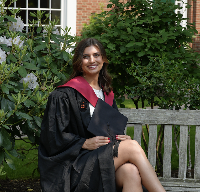 A young woman in a graduation gown sitting on a wooden bench outdoors, holding a graduation cap, surrounded by green bushes and flowering plants.