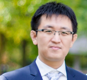 Portrait of a young man with glasses, wearing a dark suit and tie, outdoors with green trees in the background.