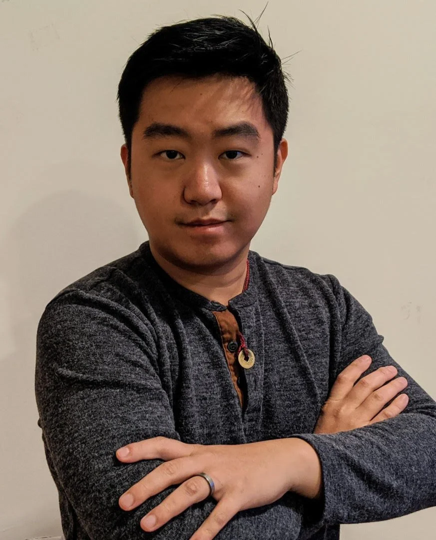 Young man with crossed arms wearing a dark gray sweater, standing against a plain beige wall.