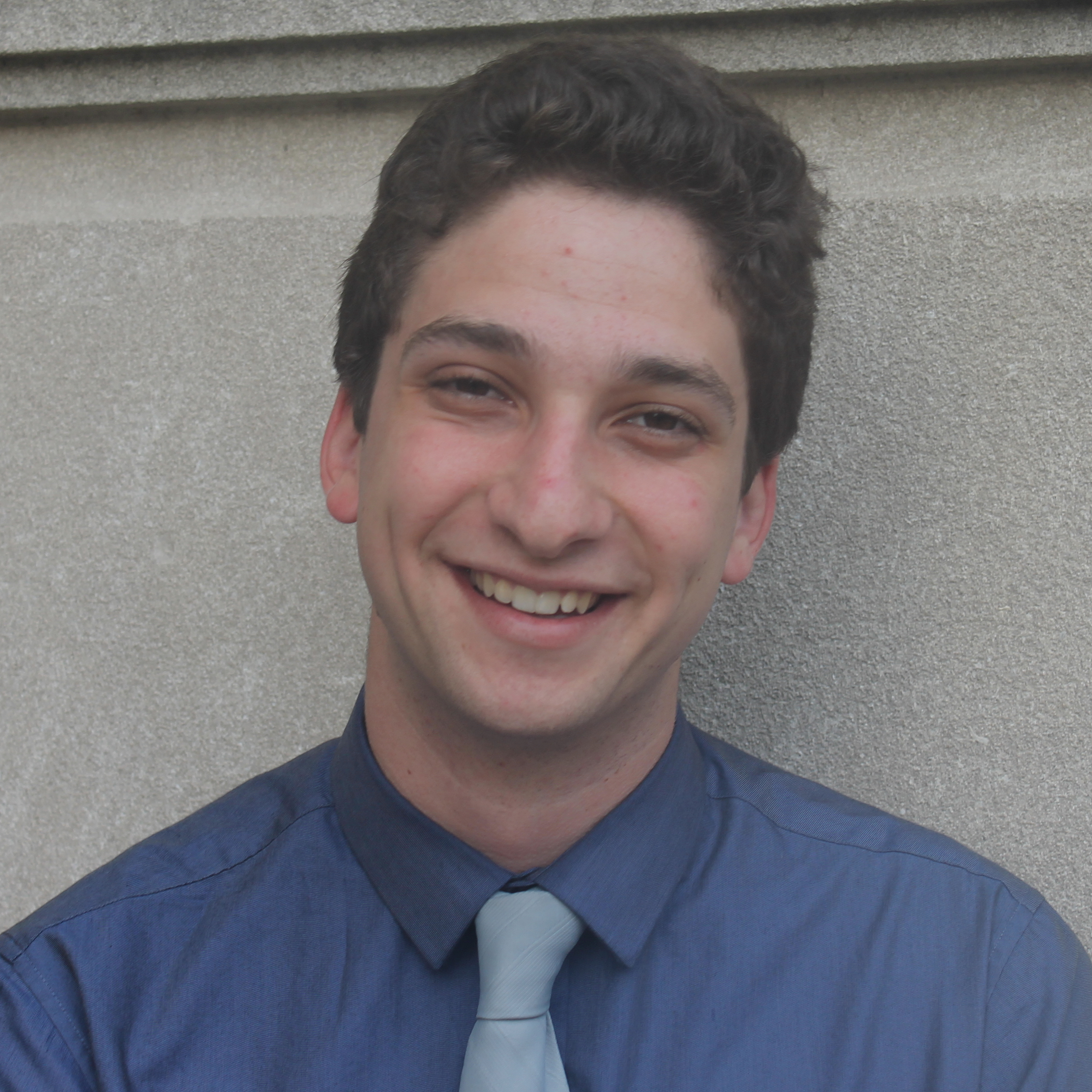 A young man with short, dark curly hair smiling, wearing a blue button-up shirt and a light gray tie, standing against a gray concrete wall.