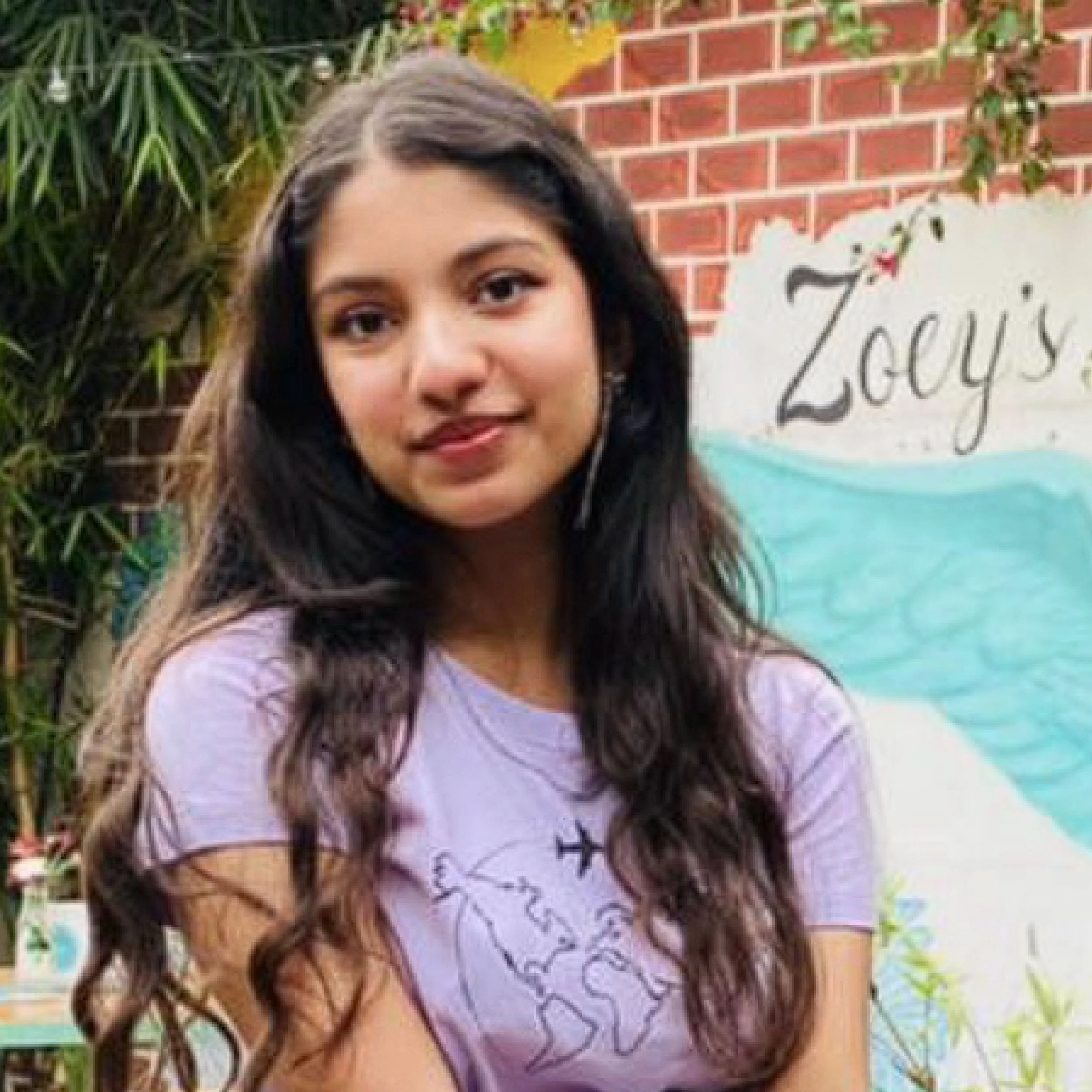 A young woman with long wavy dark hair and light brown skin smiling outdoors, standing in front of a painted sign with the text "Zoy's" and a brick wall, surrounded by greenery.