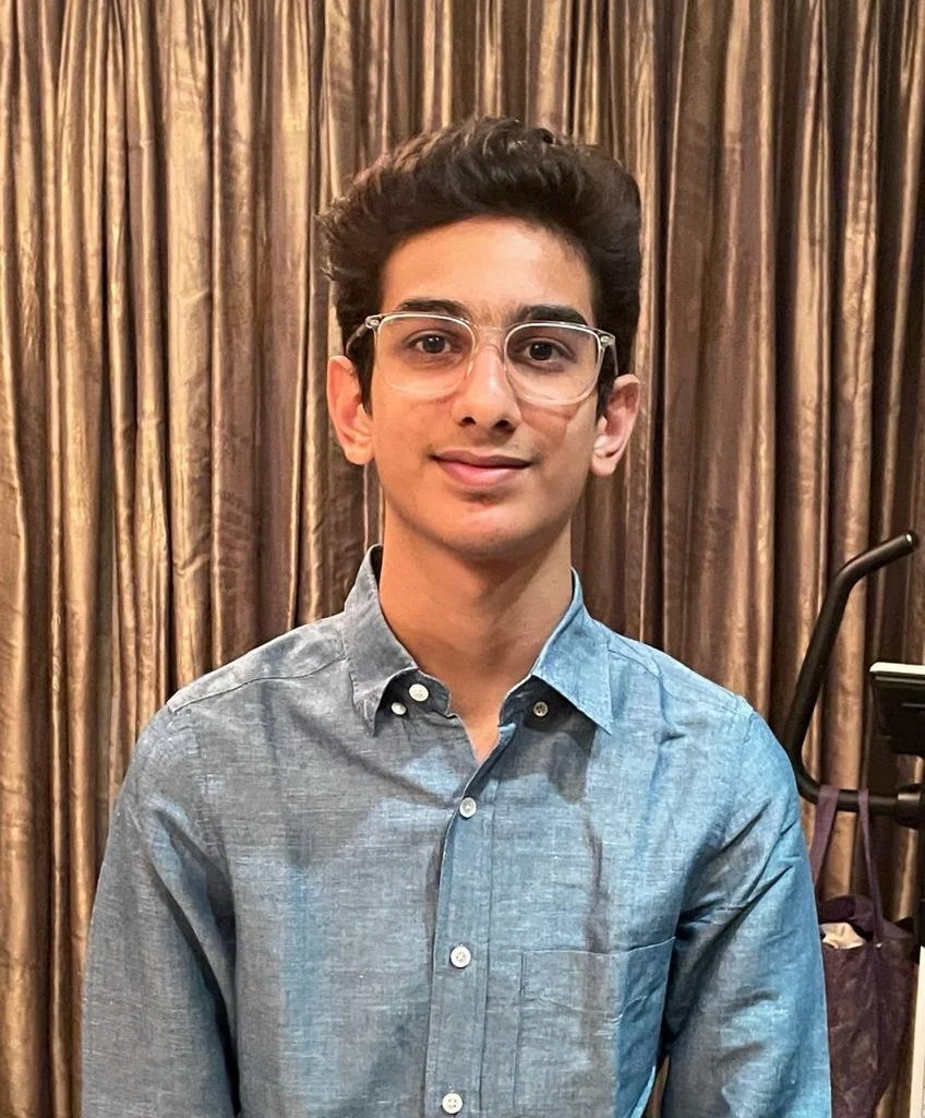 A young man with dark, curly hair, wearing clear glasses and a blue button-up shirt, standing in front of gold-colored curtains.
