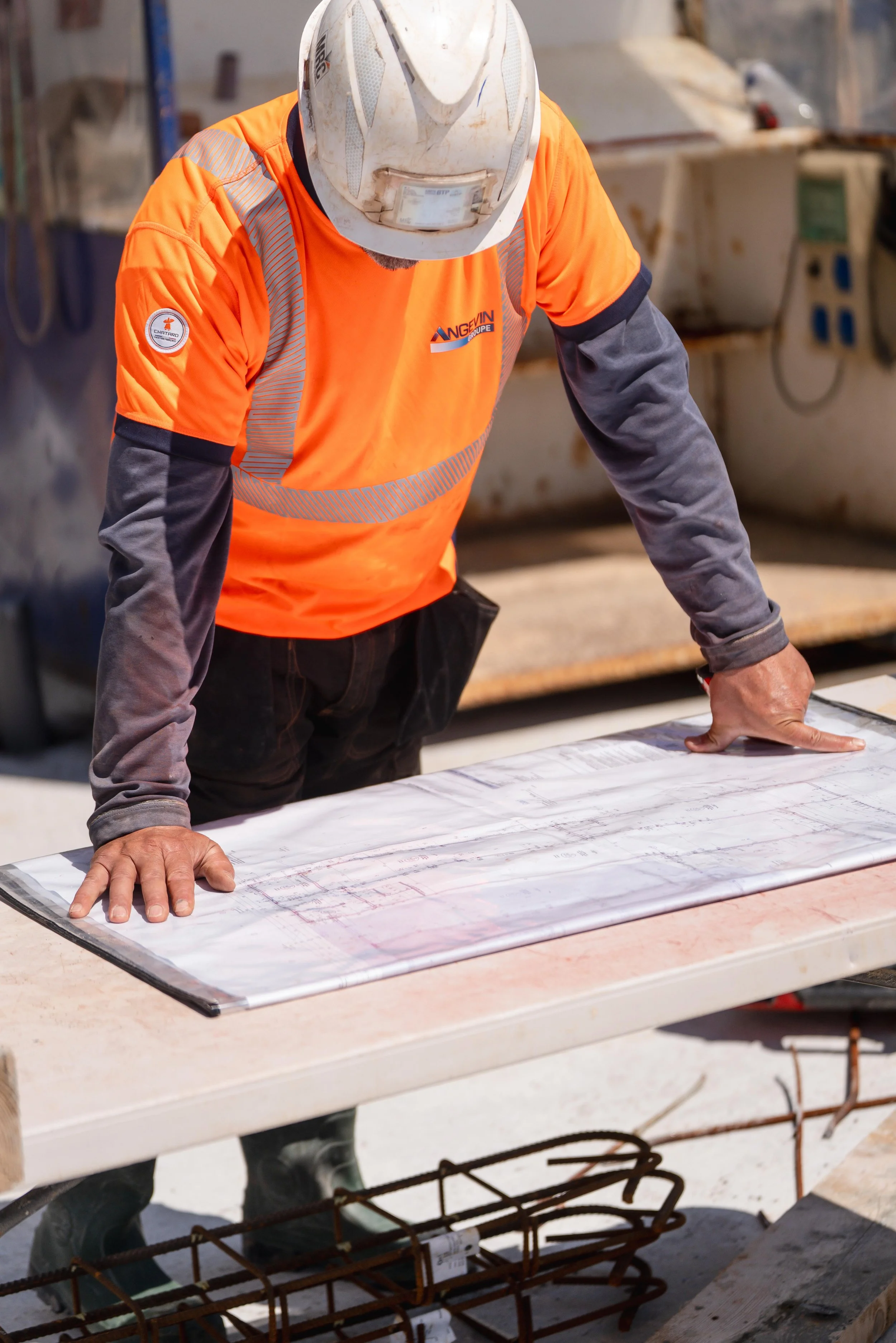 Un ouvrier portant un casque de sécurité et un gilet orange examine des plans sur une table de chantier.