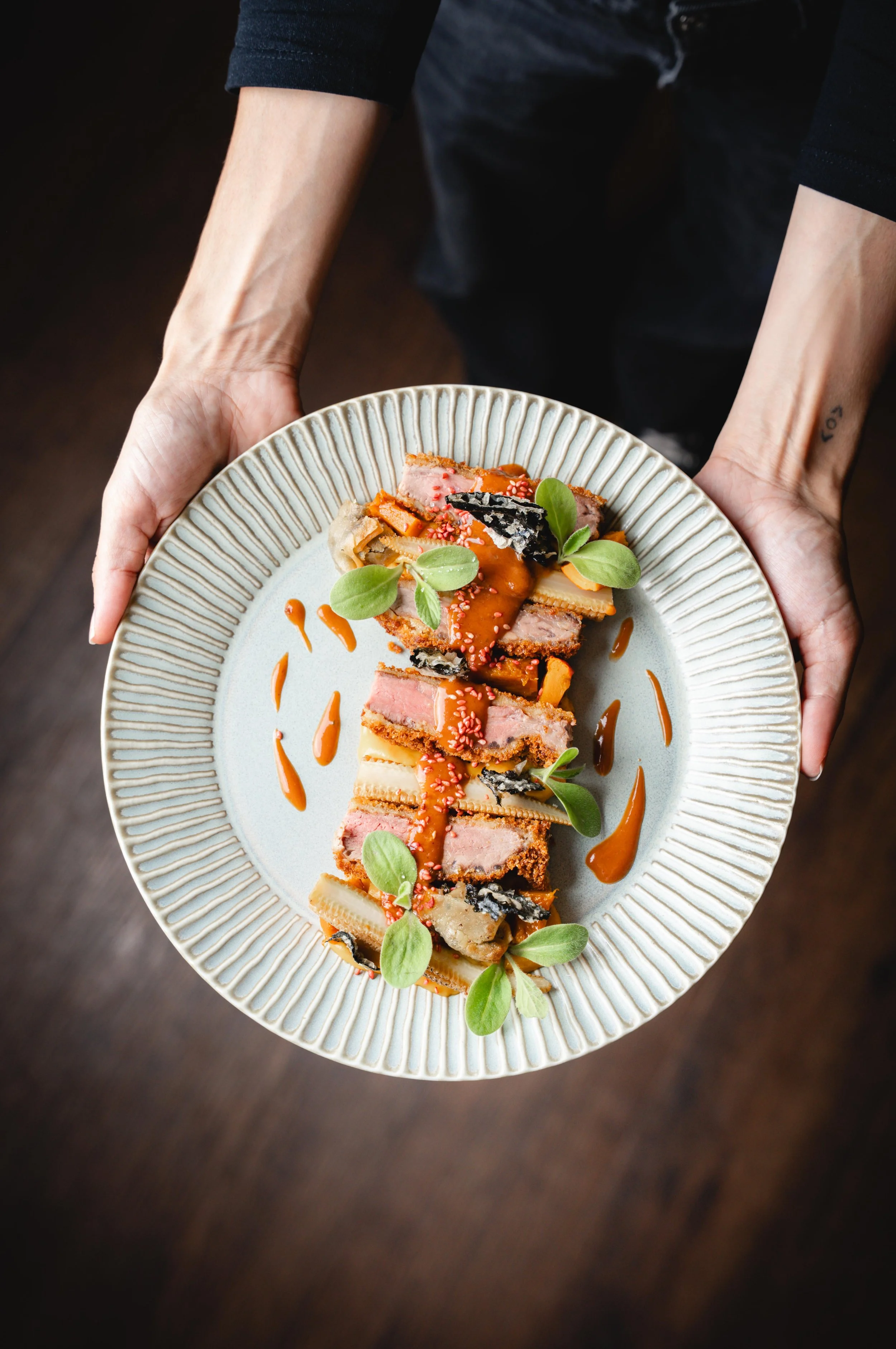 Une personne tient une assiette présentant un plat de cuisine présenté avec des sauces, des herbes vertes, et des morceaux de viande en croûte, décoré avec des graines de sésame.