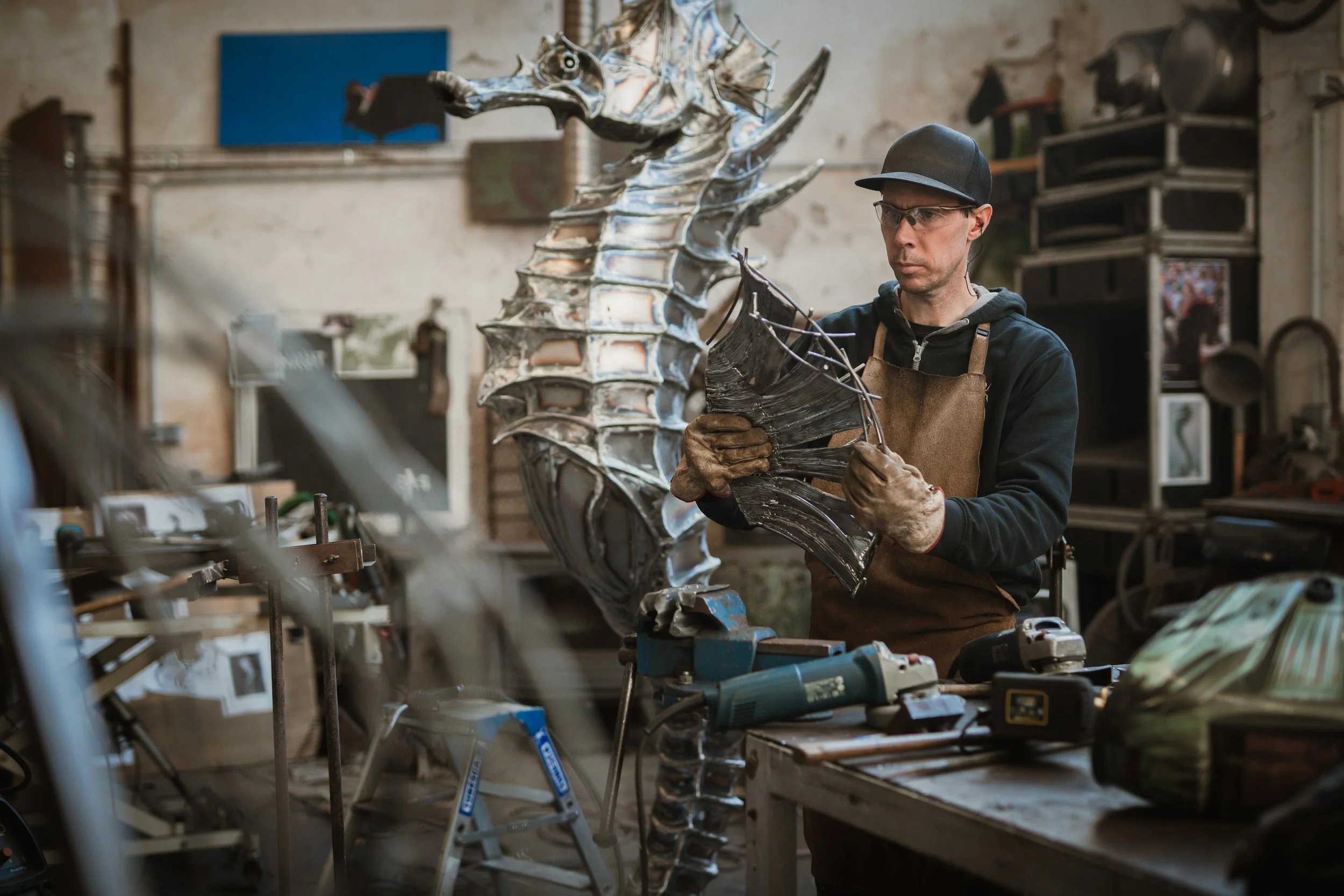 Un homme travaillant dans un atelier, façonnant une sculpture en métal représentant un dragon. Il porte des lunettes de sécurité, un casque noir, un tablier en cuir et des gants.