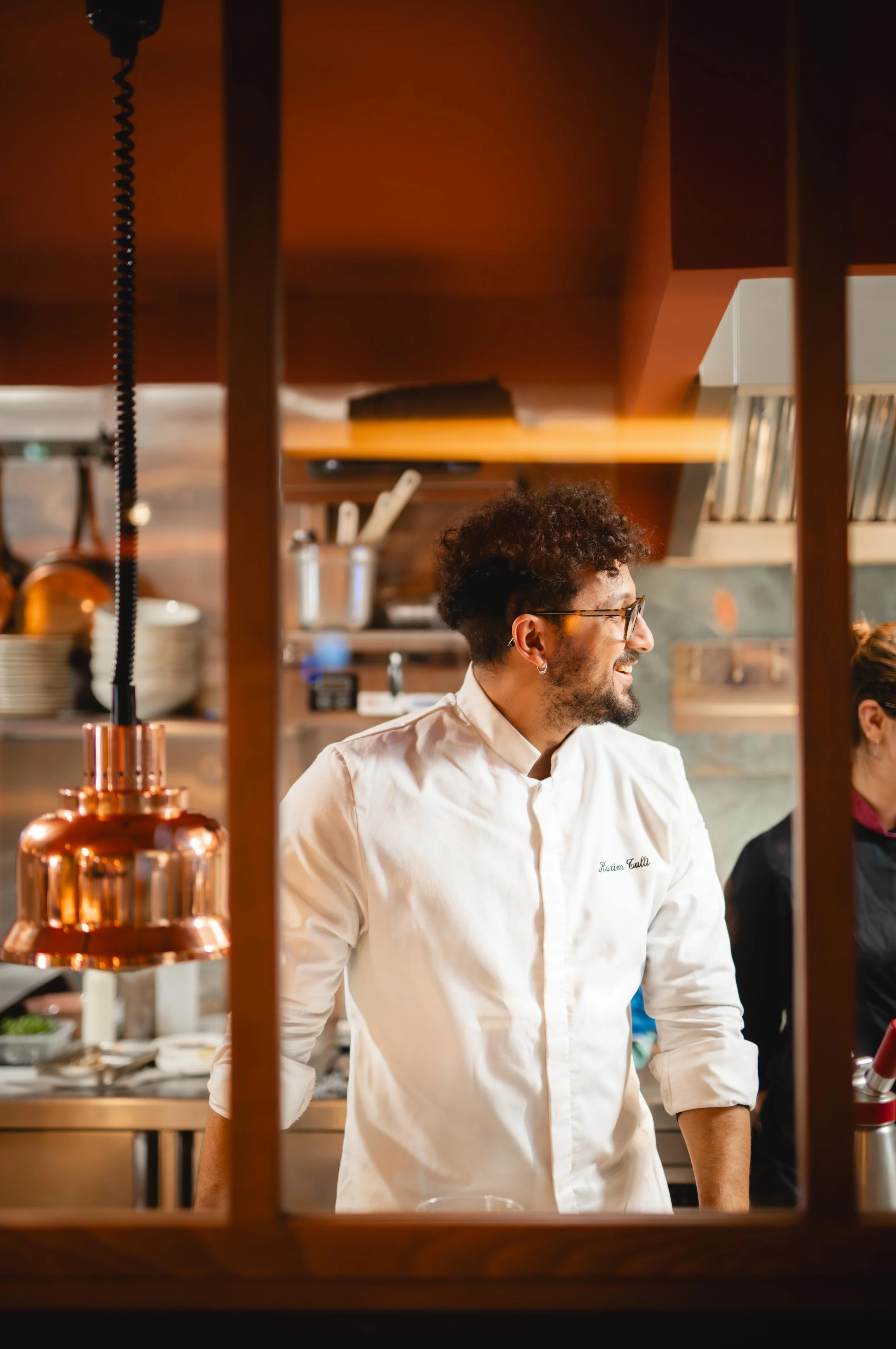 Un chef souriant dans une cuisine ouverte, portant une veste blanche, avec un fond de cuisine en bois, des ustensiles et des plats.