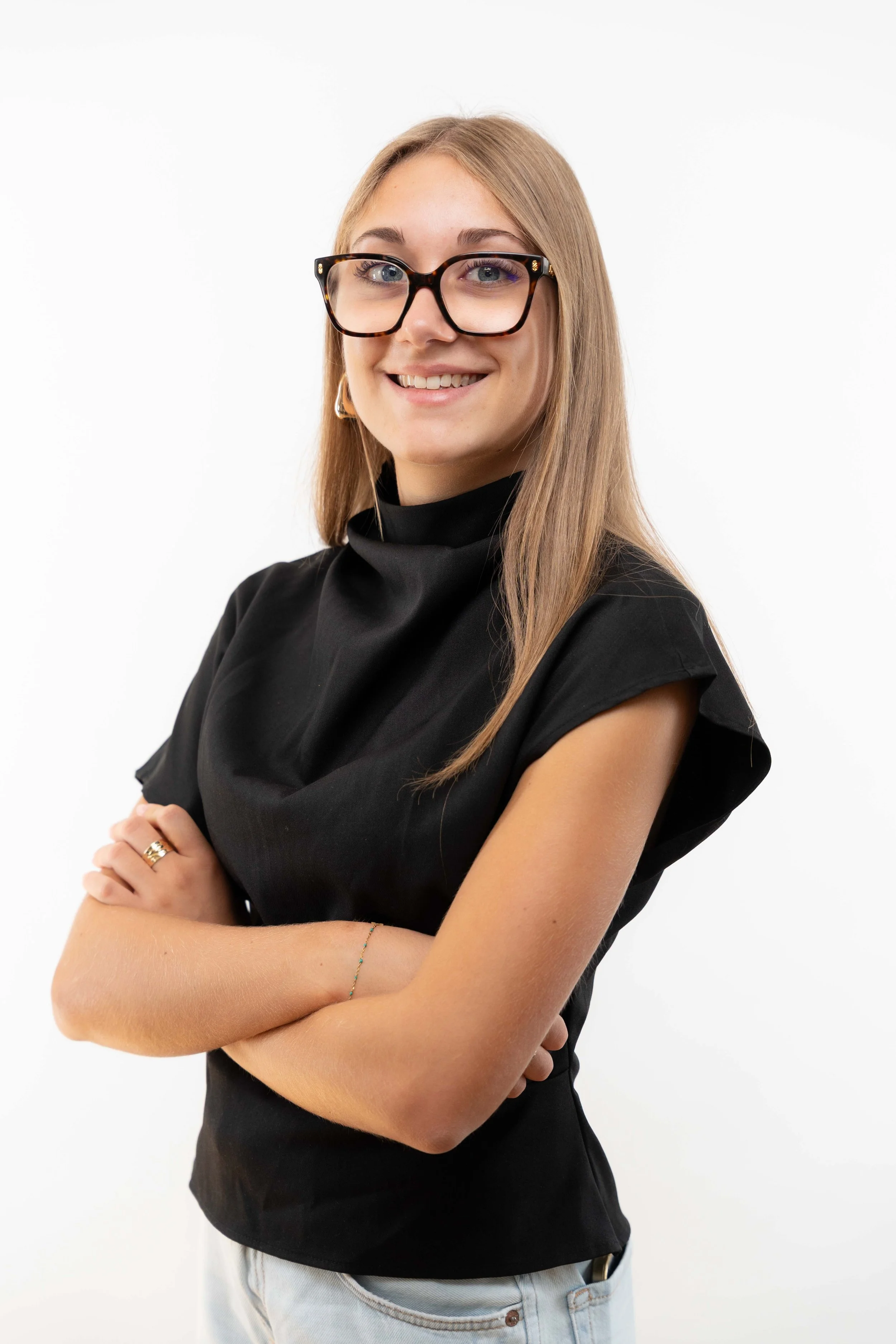 Jeune femme avec lunettes à la monture tortoise, porte un haut noir à col roulé, bras croisés, sourire, cheveux longs et lisses, fond blanc.