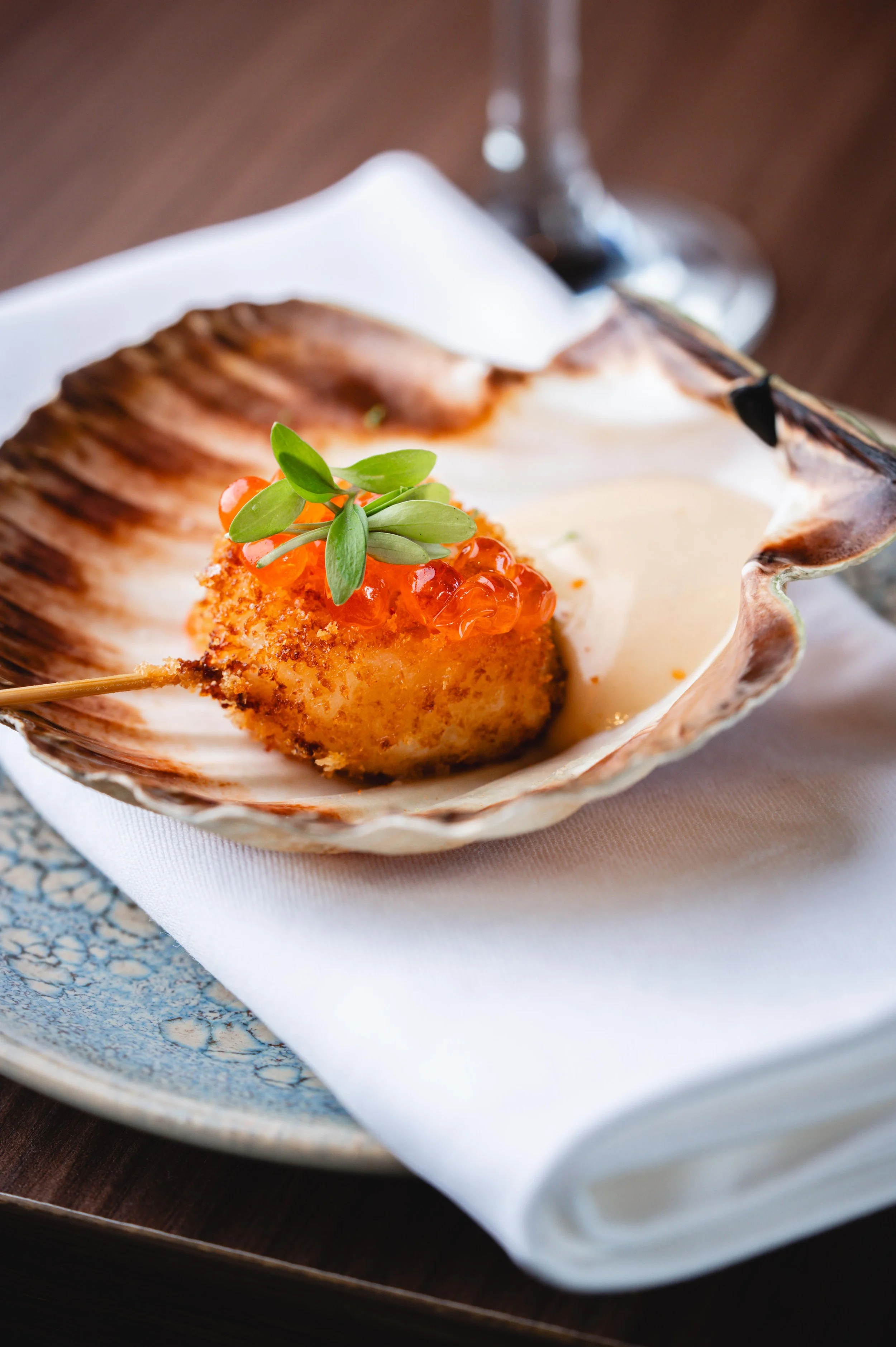 Un plat de fruits de mer présenté dans une coquille de scallop, garnie de petite plante verte, avec une sauce blanche et un verre de vin en arrière-plan.
