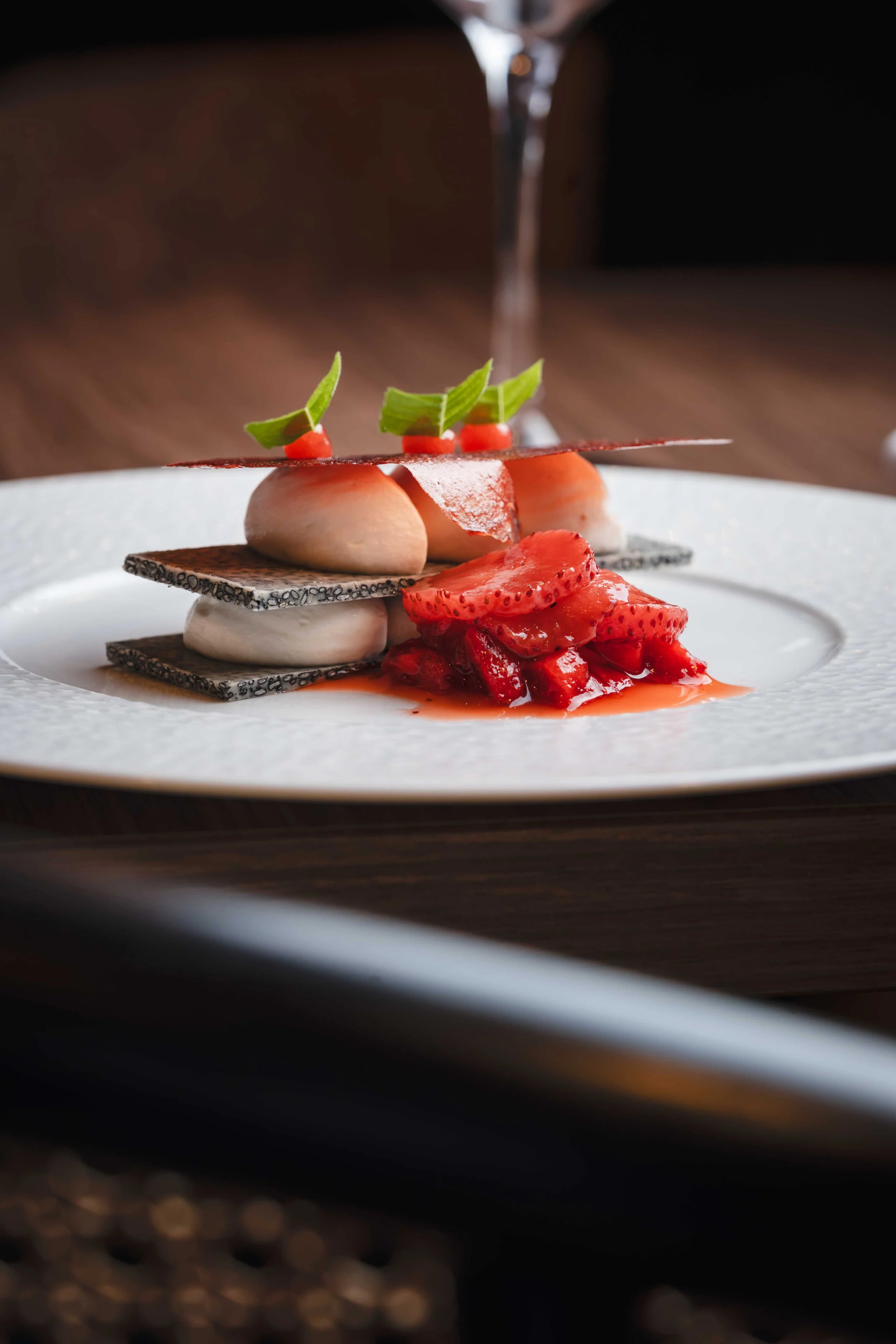 Un dessert sophistiqué avec des fraises, des feuilles vertes, des couches de biscuits et une sauce rouge, servi sur une assiette blanche, avec un verre de boisson en arrière-plan.