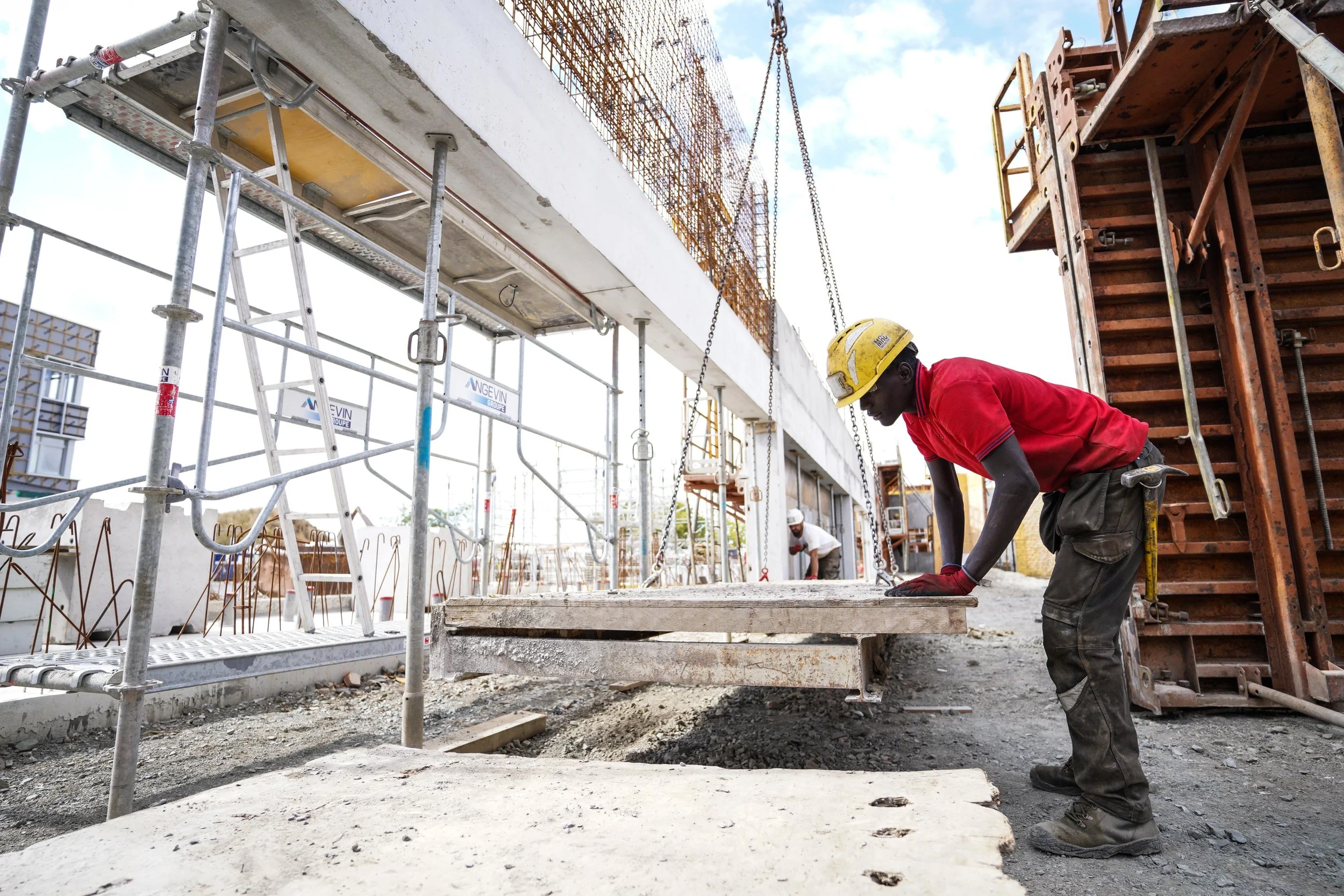 Un ouvrier en casque de construction travaille sur un chantier, pliant ou ajustant une pièce de béton, avec des structures métalliques et un échafaudage autour de lui.