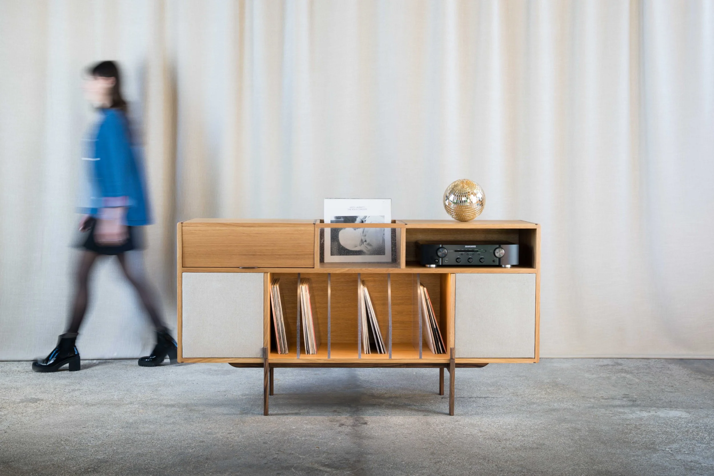 Une personne marche rapidement à côté d'une étagère en bois contenant des disques vinyles, un cadre photo, une décoration en forme de boule disco et un lecteur audio. Le fond est un mur beige avec des rideaux.