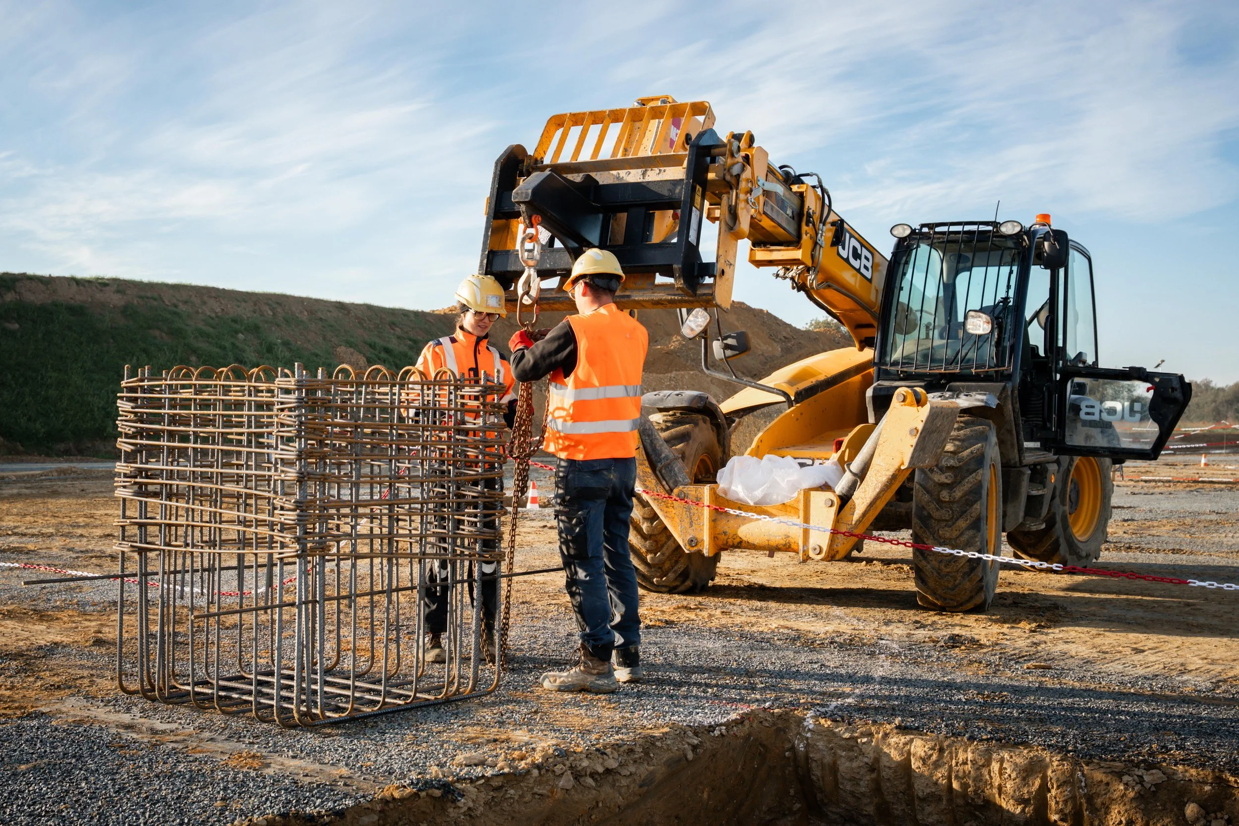 Deux ouvriers travaillant sur un chantier de construction avec une grue JCB équipée d'une pince mécanique, en train de manipuler des armatures en acier pour le renforcement du béton, sous un ciel partiellement nuageux.