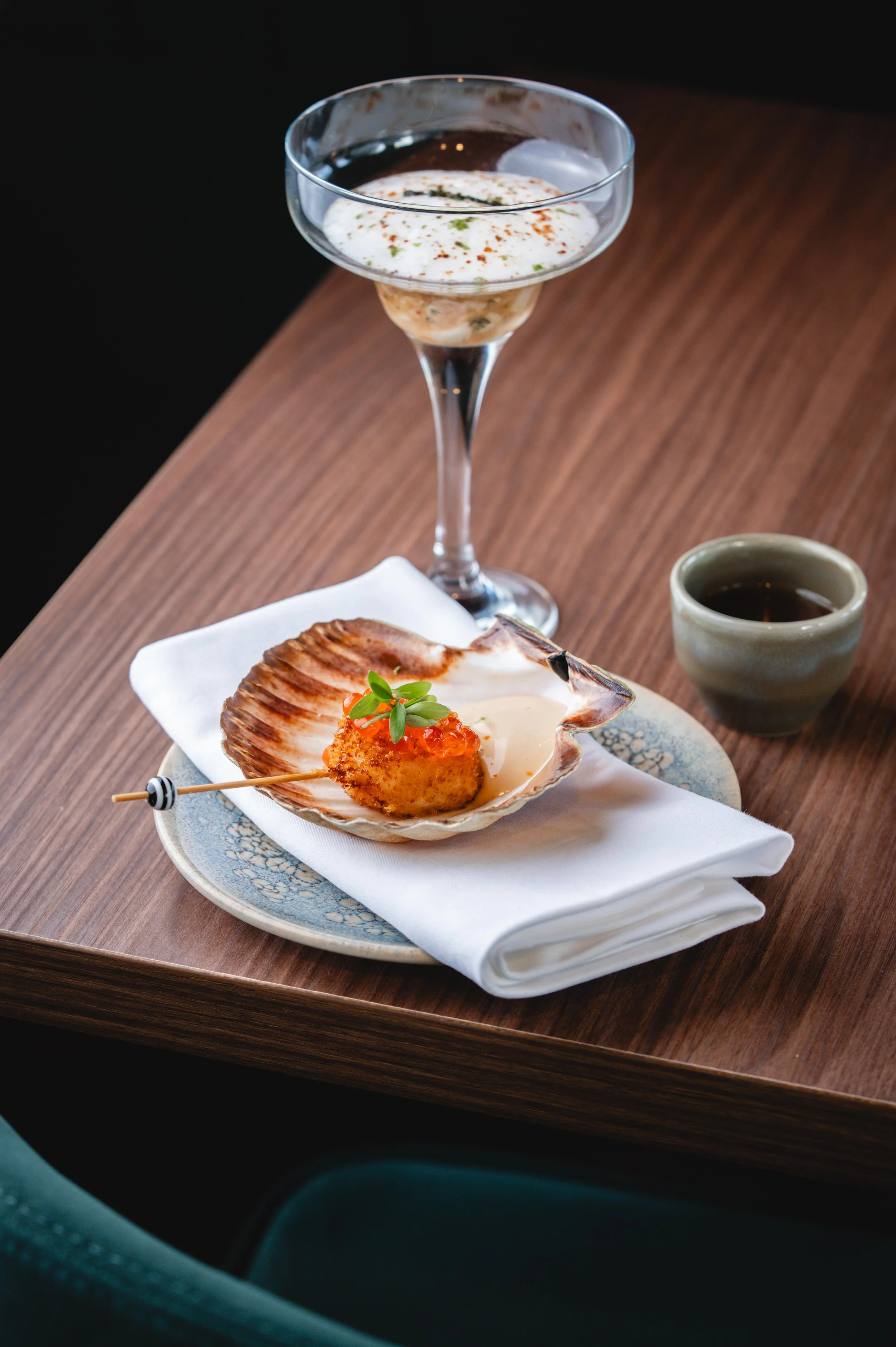 Huitres garnies de saumon, oeufs de saumon et microgreens, servies sur une assiette blanche avec un napperon en tissu blanc, accompagnées d'une coupe de cocktail à la crème et d'une tasse de thé ou café, sur une table en bois.