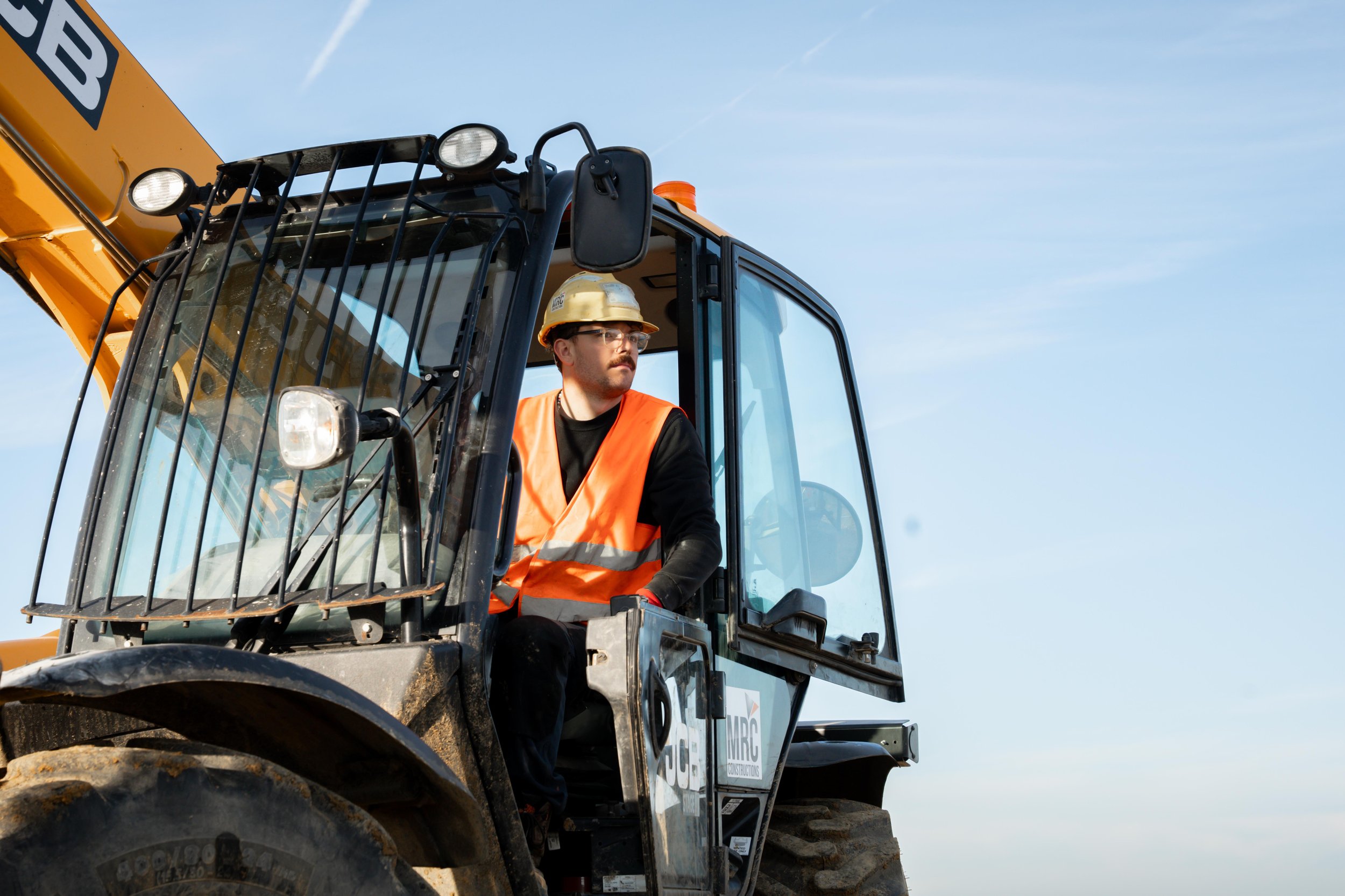 Un homme portant un casque et un gilet de sécurité orange, assis dans la cabine d'une machine de construction, près d'une pelle mécanique, sous un ciel clair.