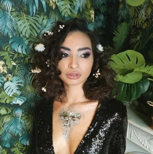 This is a portait of a curly haired woman who is standing against a background of tropical green leaves. Her hair is collarbone length and she is wearing a black sequinned v-neck top. She has white flowers pinned throughout her hair.
