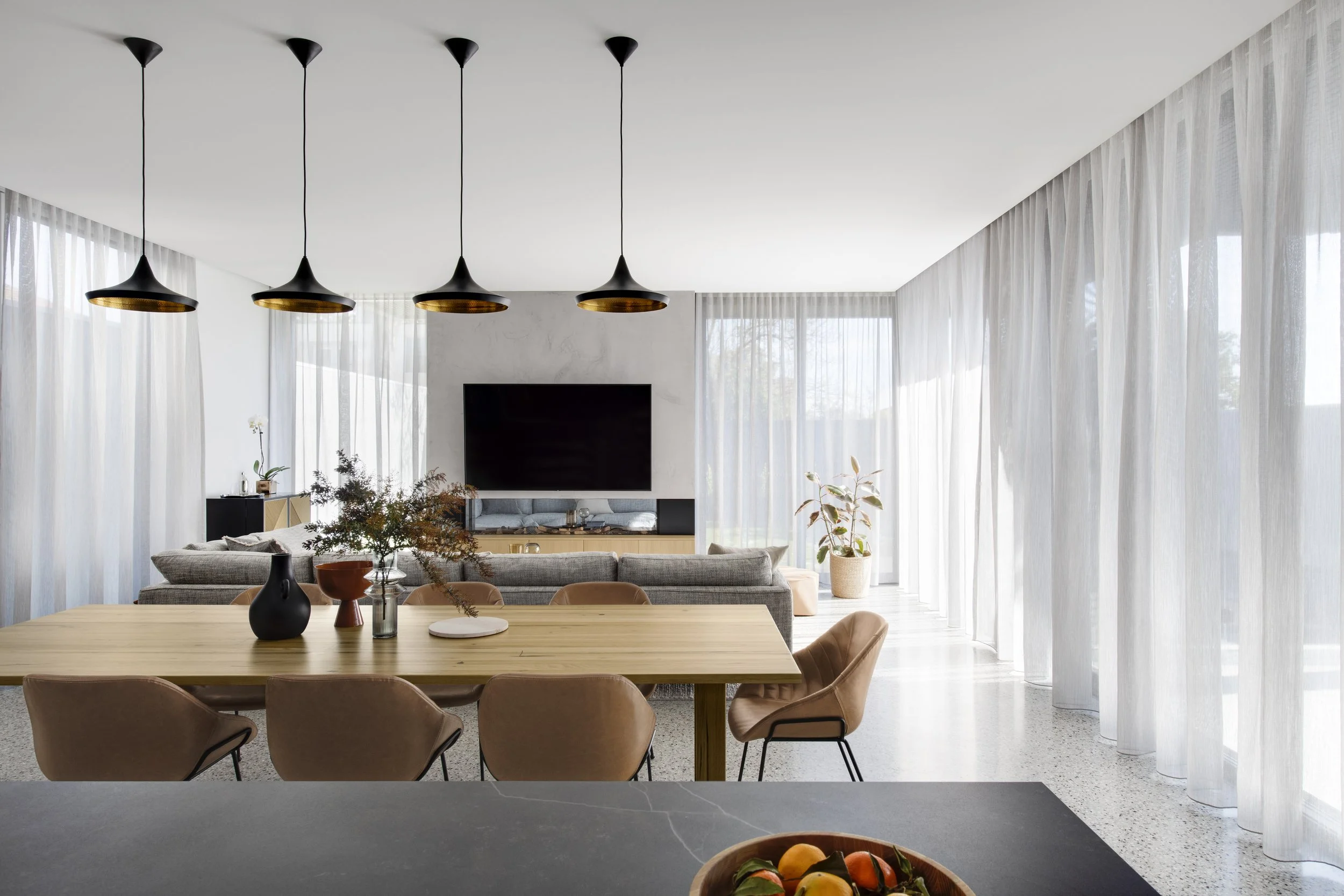 Modern living and dining room with large windows and sheer curtains, a wooden dining table with beige chairs, pendant lights over the table, a gray sofa, a large flat-screen TV, potted plants, and decorative vases.
