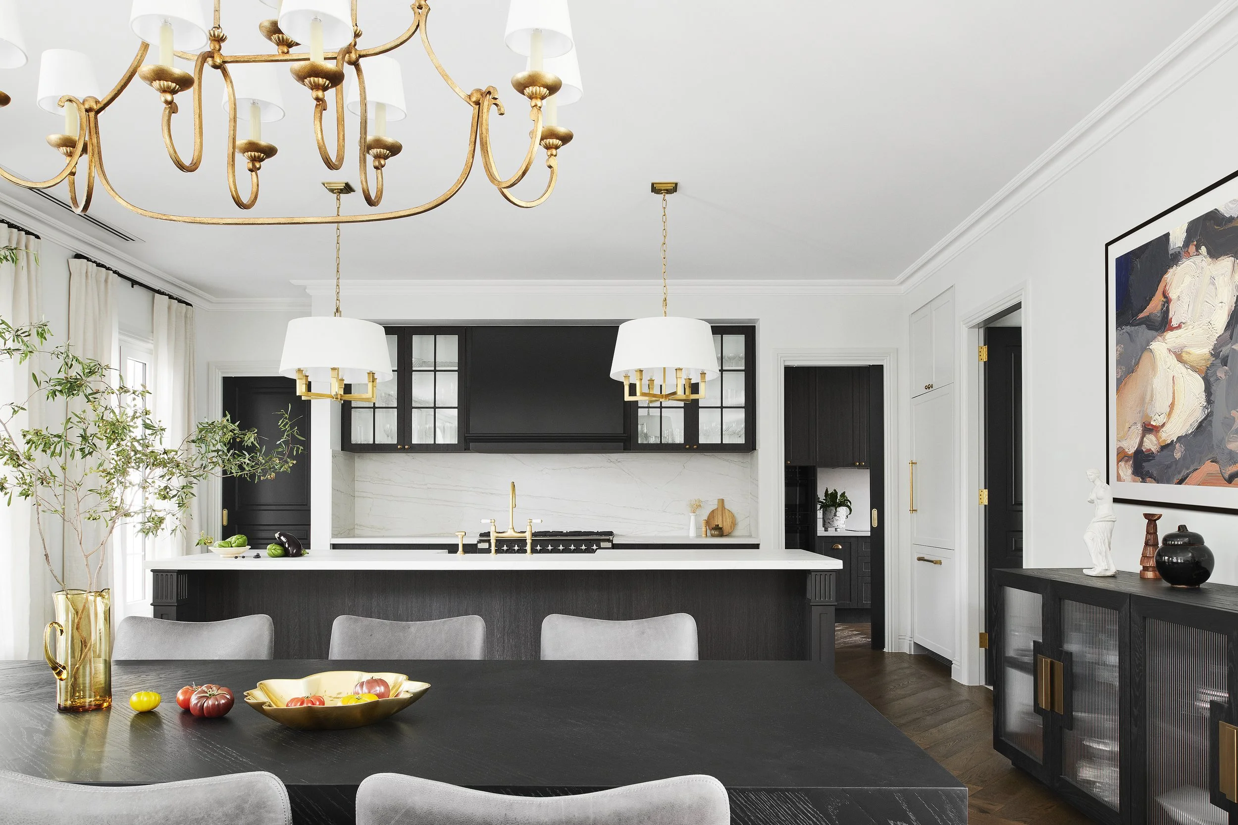 Modern kitchen with black and white cabinetry, including a large black island with a white marble countertop, gold fixtures, and two black pendant lights with white shades hanging above. A dining table with gray chairs is in the foreground, decorated with a gold bowl of tomatoes, along with a black sideboard displaying decorative statues and vases, and a large abstract painting on the wall.