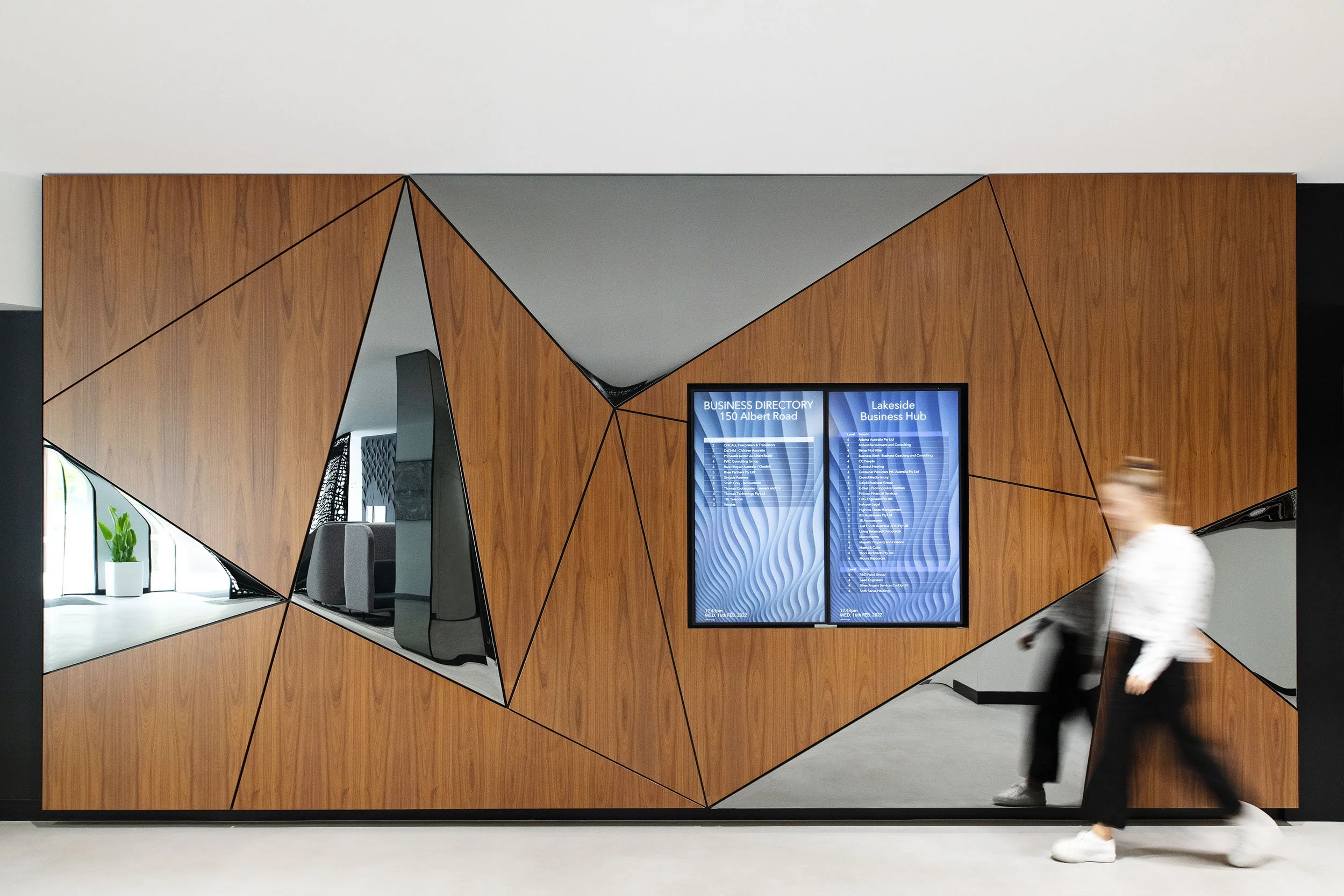 Modern interior with a wooden wall featuring geometric cutouts, a digital directory sign, a person walking by, and a window with a plant on the sill.