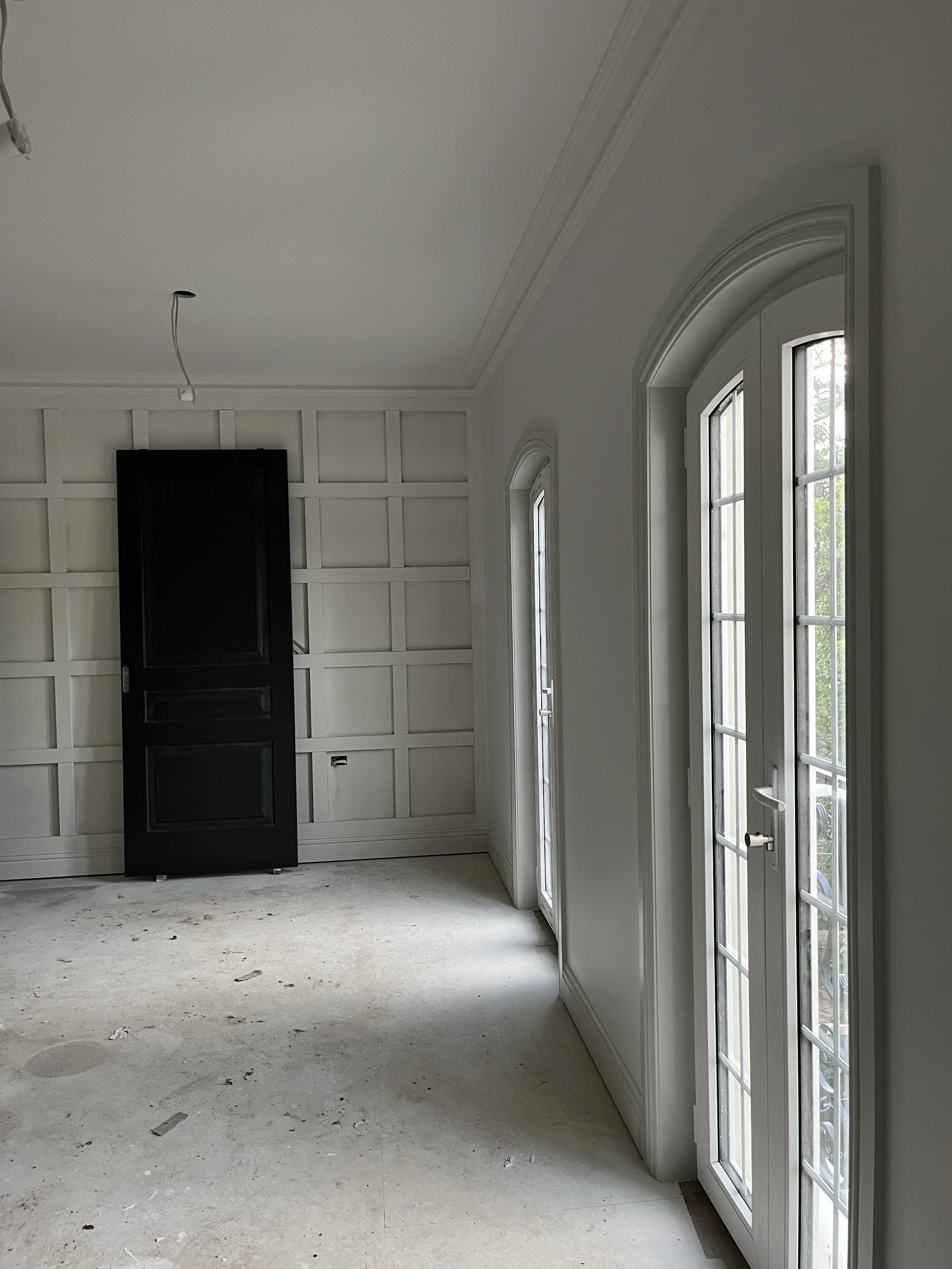 Empty room with white walls and large arched windows with grid pattern, leading to a balcony or outdoor space, with a black door or cabinet leaning against the wall, and an unfinished floor.