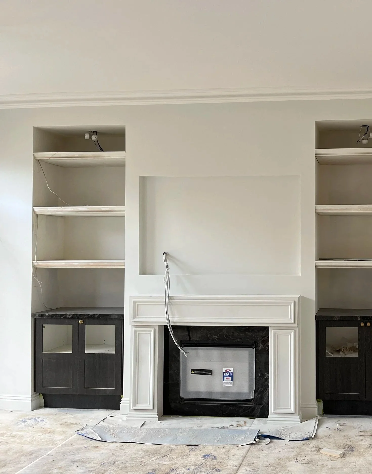 Living room with built-in shelves, a fireplace with black marble surround, and unfinished wiring and cabinetry in a stylish home.