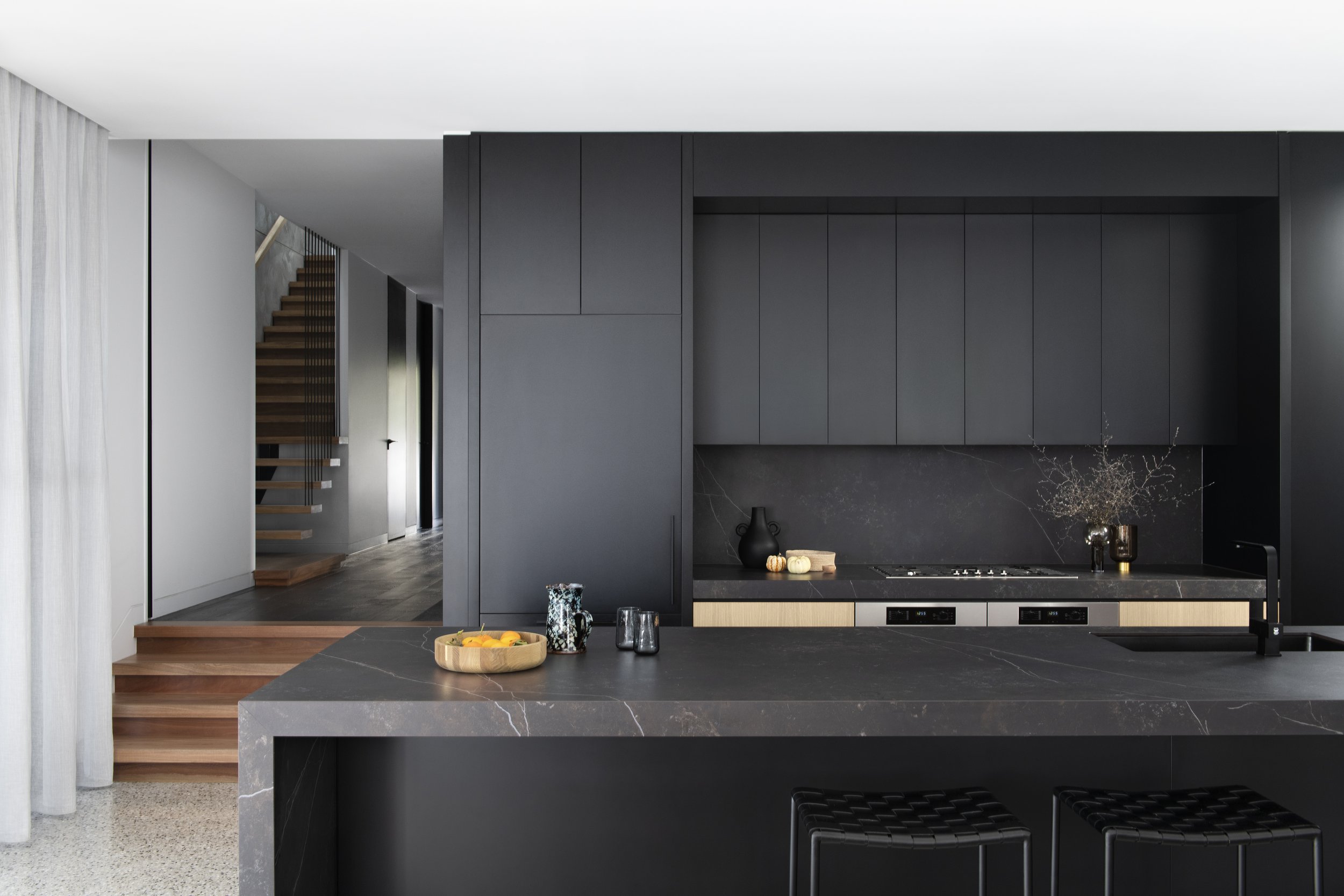 Modern kitchen with black cabinets, marble countertop island, and minimal decor including black and gold vases, a wooden fruit bowl, and small pumpkins. Wooden stairs are visible in the background.