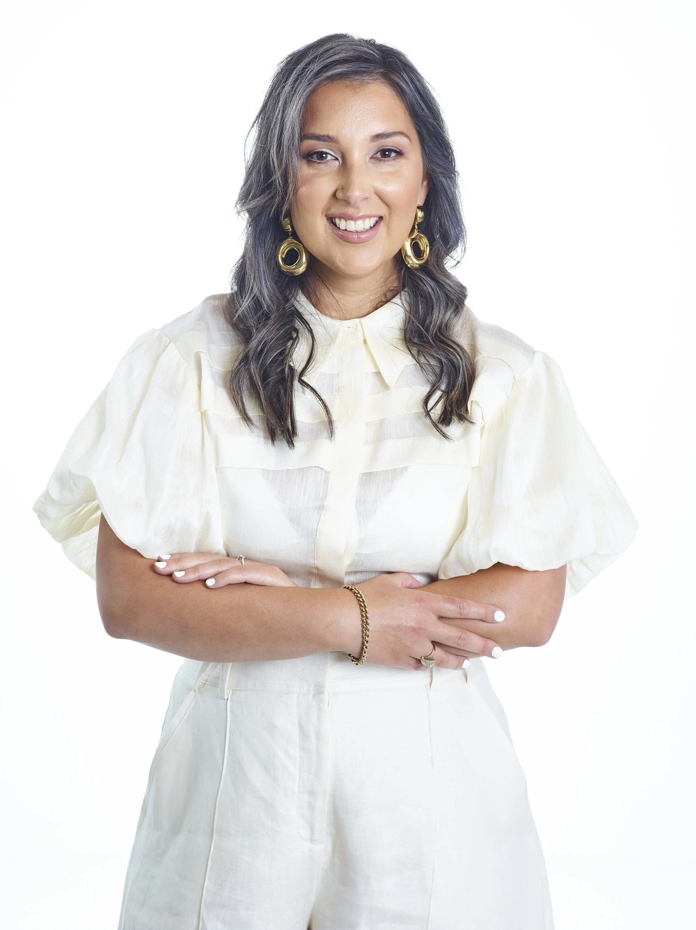 A woman with dark wavy hair, wearing a cream-colored blouse with puff sleeves, Gold hoop earrings, a bracelet, and a ring, standing with arms crossed and smiling against a plain white background.