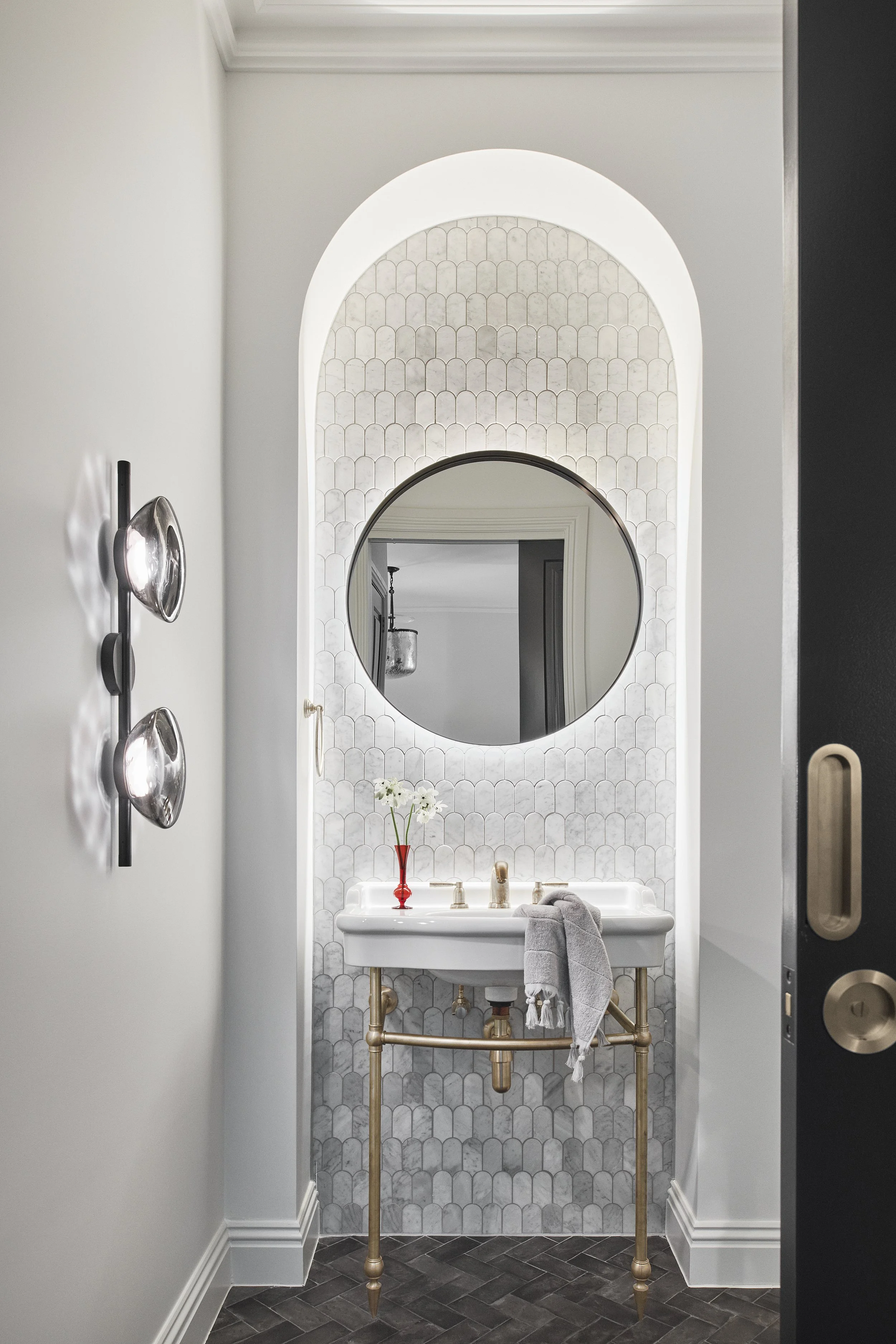 A small bathroom with a white sink on a gold-colored stand, a round mirror above it, and a red vase with white flowers. The wall behind has a light, patterned tile design, and there is a gray towel hanging on the sink.