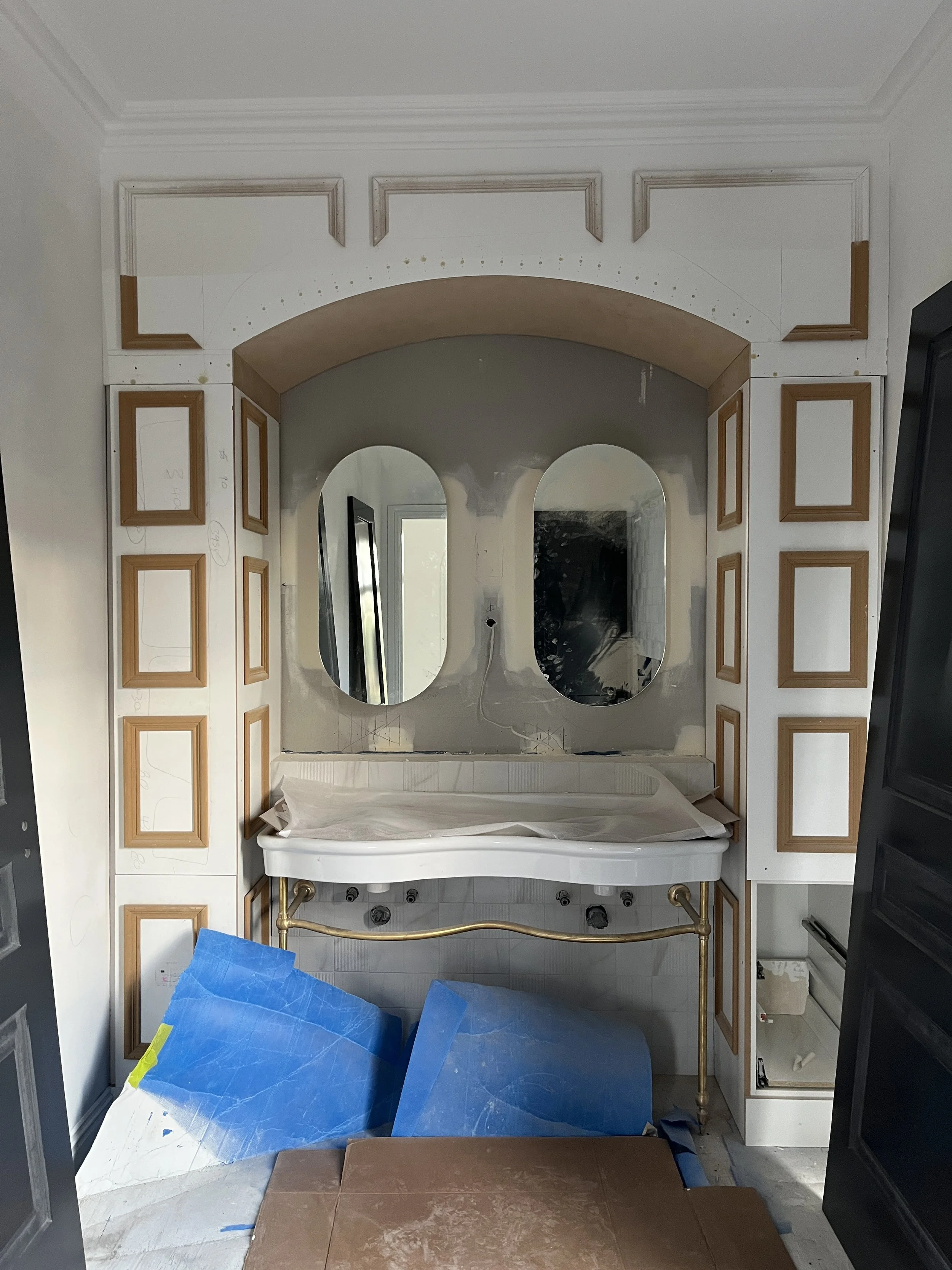 Bathroom vanity area under construction with two oval mirrors, an ornate gold-colored faucet, and wooden framed panels on the wall. Construction materials and protection coverings are present.