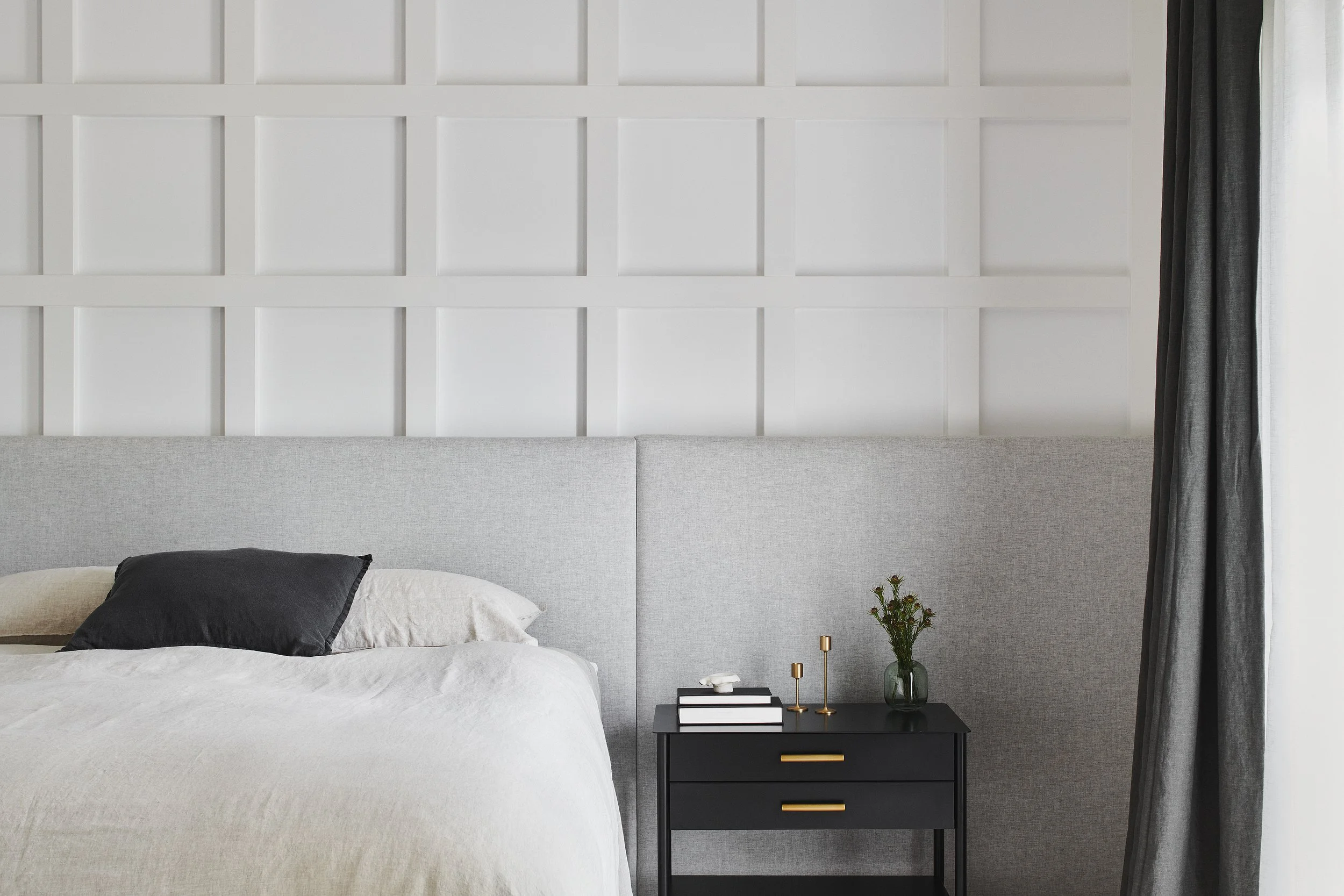 A neatly made bedroom with a gray headboard, white bedding, and pillows, next to a black nightstand with gold handles, decorated with a small green plant, candles, and a stack of books, next to a window with white and gray curtains.