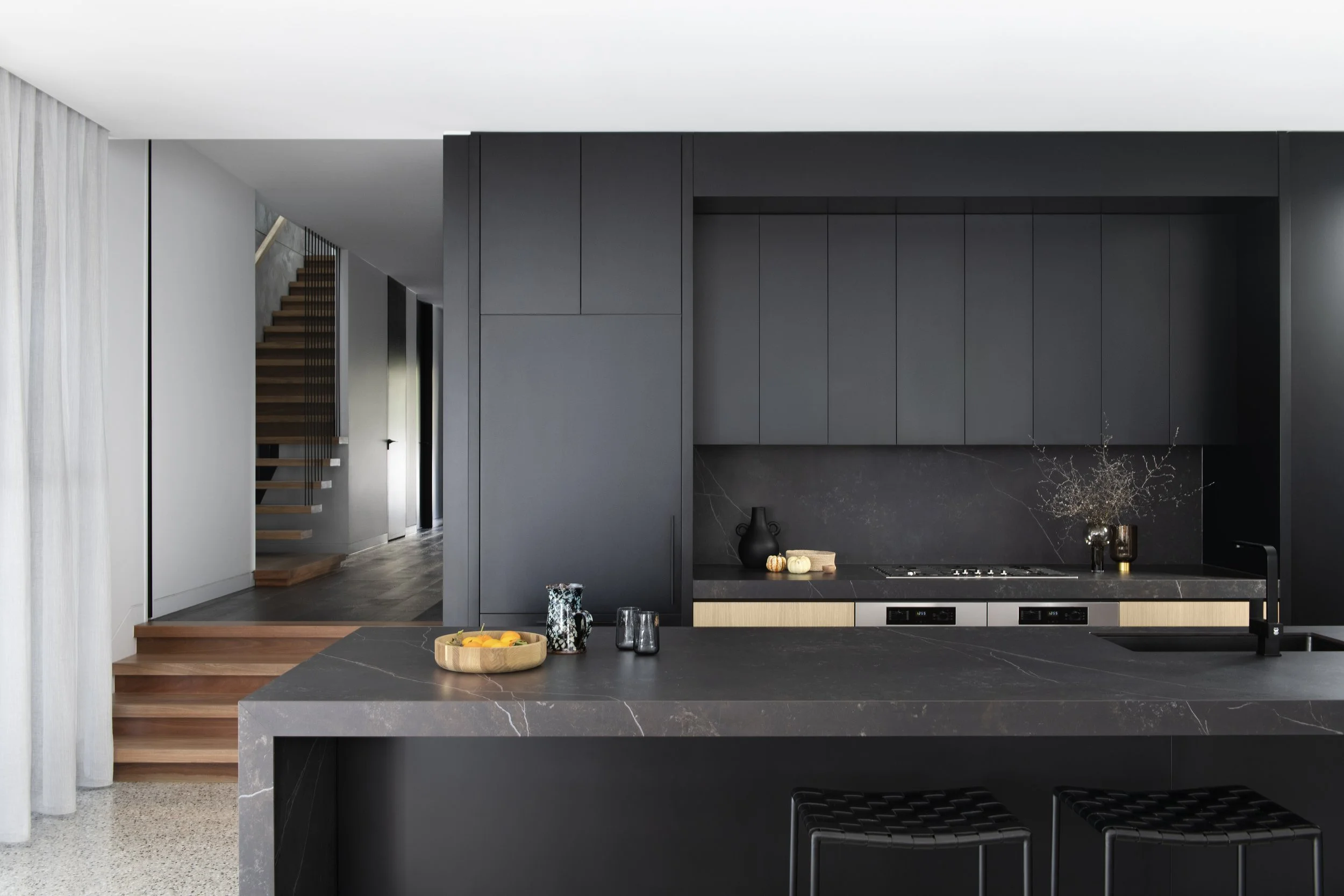 Modern kitchen with black cabinets and a black marble backsplash, a large black marble kitchen island with a wooden bowl of oranges, and a view of a floating wooden staircase in the background.