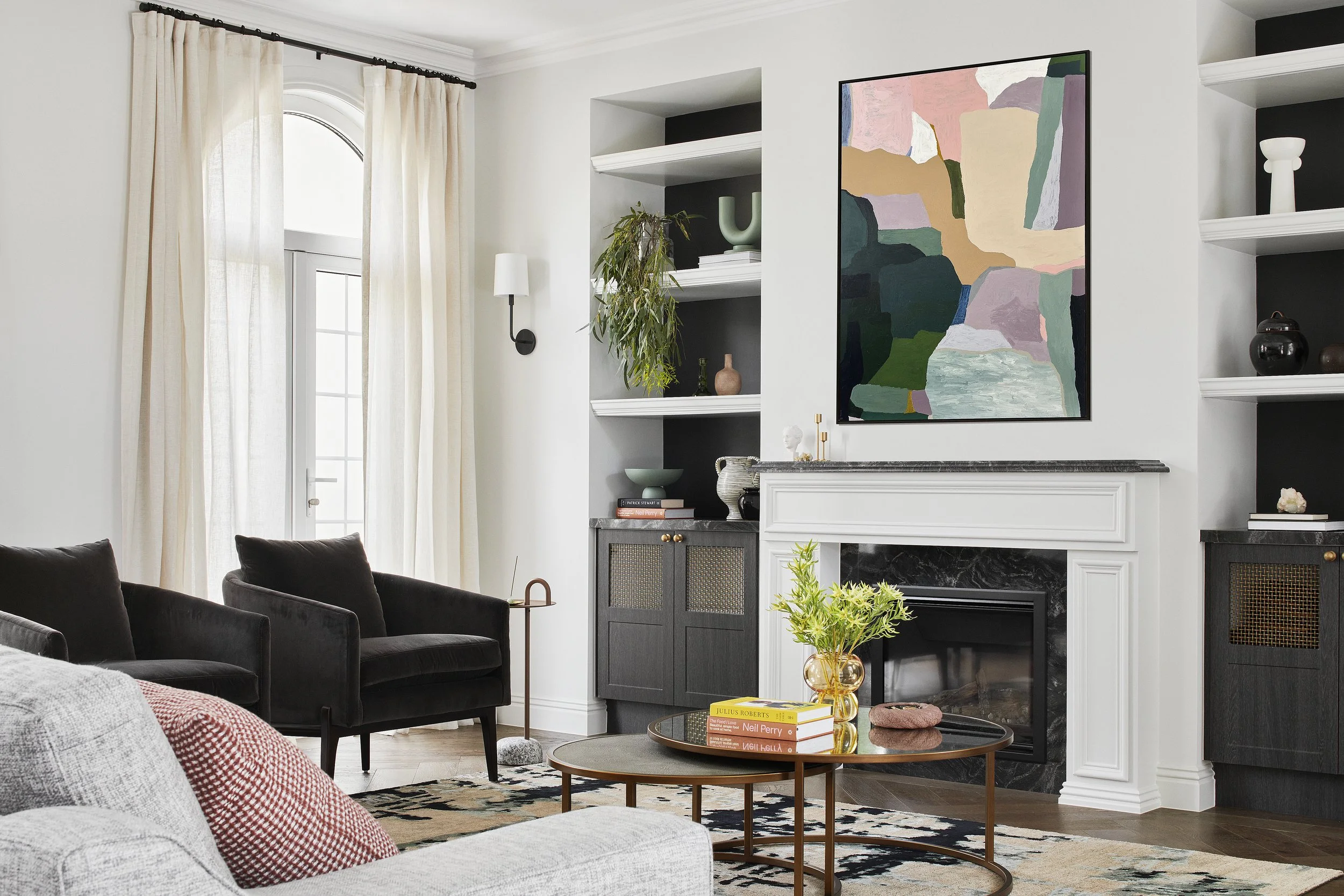 Living room with white walls, beige curtains, a large window, a black armchair, a white couch with a red pillow, a round coffee table with books and a vase of greenery, a white fireplace with black marble trim, and wall shelves with decorative objects, a colorful abstract painting above the fireplace.