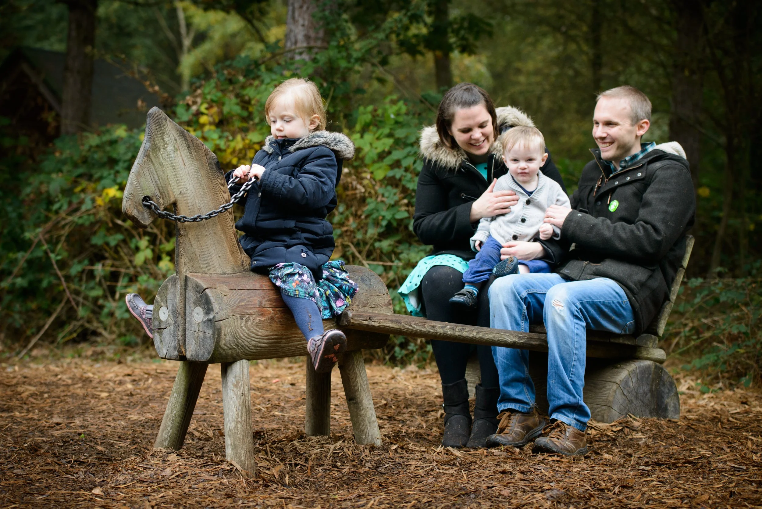 A family of four sitting on a wooden park bench with a horse-shaped wooden playground sculpture nearby in a wooded area. The mother and father hold two young children, one of whom is sitting on the mother's lap, while the other is sitting on the bench.