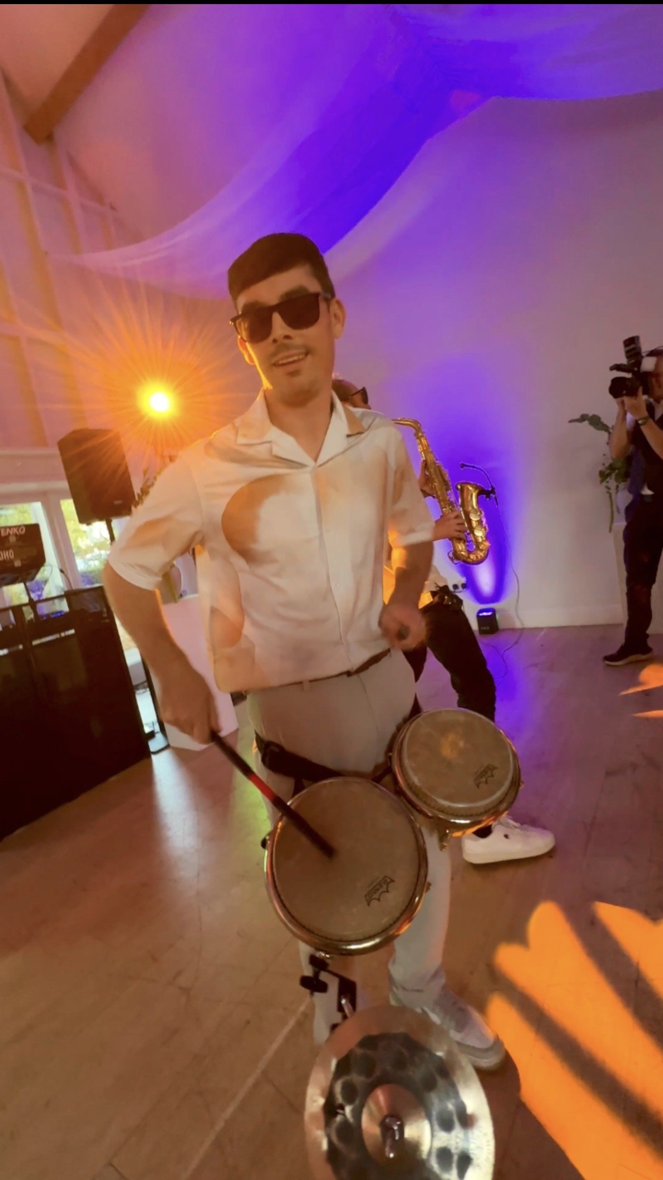 A man wearing sunglasses, a light-colored shirt, and white pants is playing the drums at a lively indoor event with colorful lighting. A saxophone player and a photographer are visible in the background.