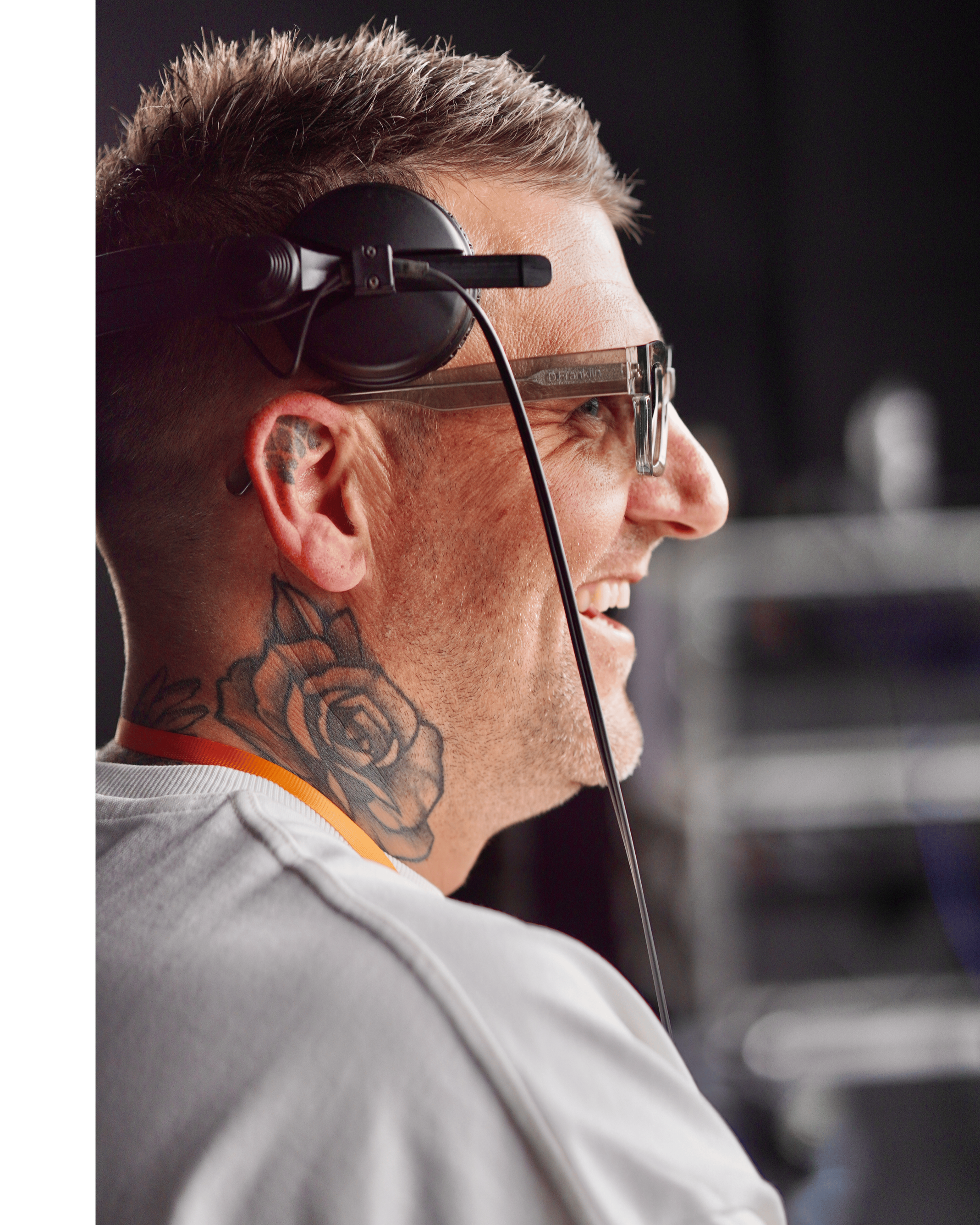 A man wearing headphones, glasses, and a white shirt, smiling and looking to the right. He has a tattoo of a rose on his neck.