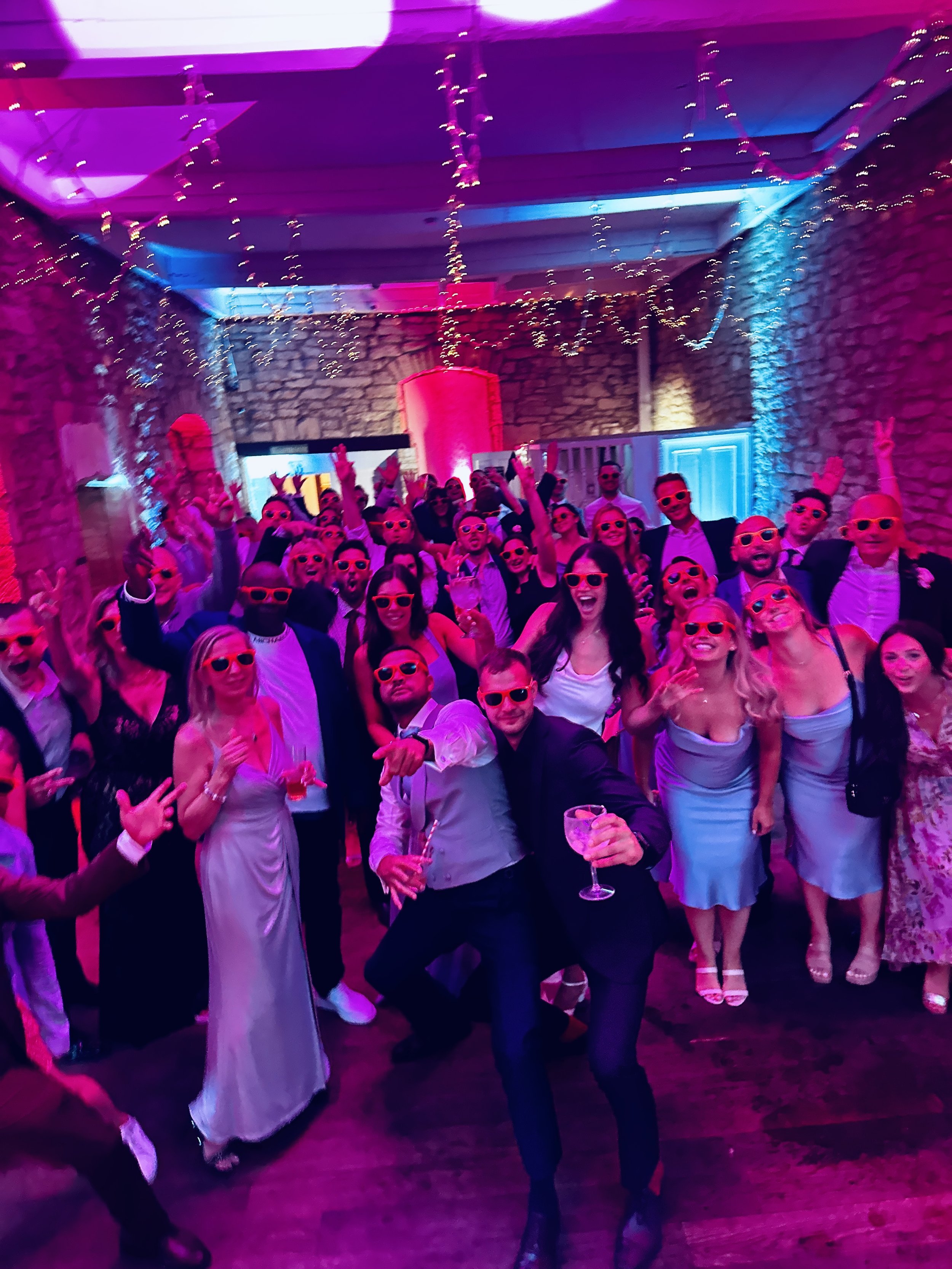 A large group of people at a lively party or celebration, many wearing pink sunglasses and dressed in formal attire, dancing and having fun under colorful lighting with string lights hanging from the ceiling.