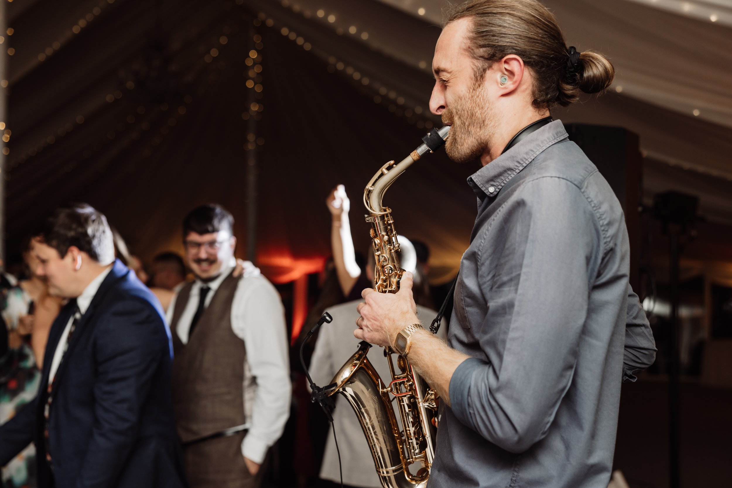 A man with long hair tied in a bun playing a saxophone at a lively indoor event, with other people dancing and enjoying the music in the background.