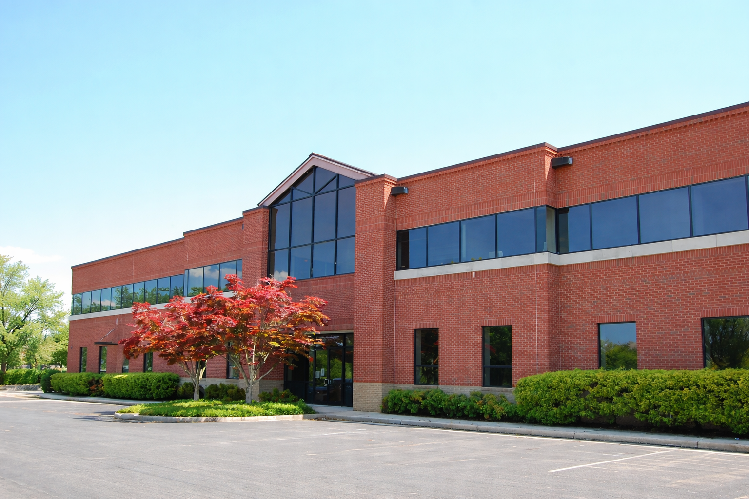 Exterior of a two-story red brick commercial building with a central glass atrium, blue-tinted windows, landscaped shrubs, and an empty parking lot under a clear sky.