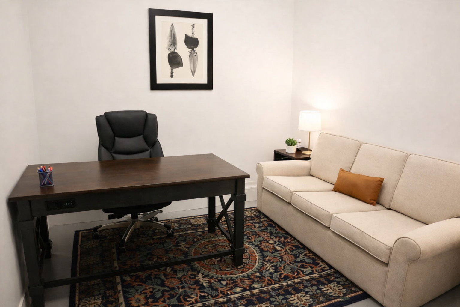 Small furnished office with a dark wood desk and chair, a beige sofa with a side table and lamp, patterned area rug, and framed wall art.